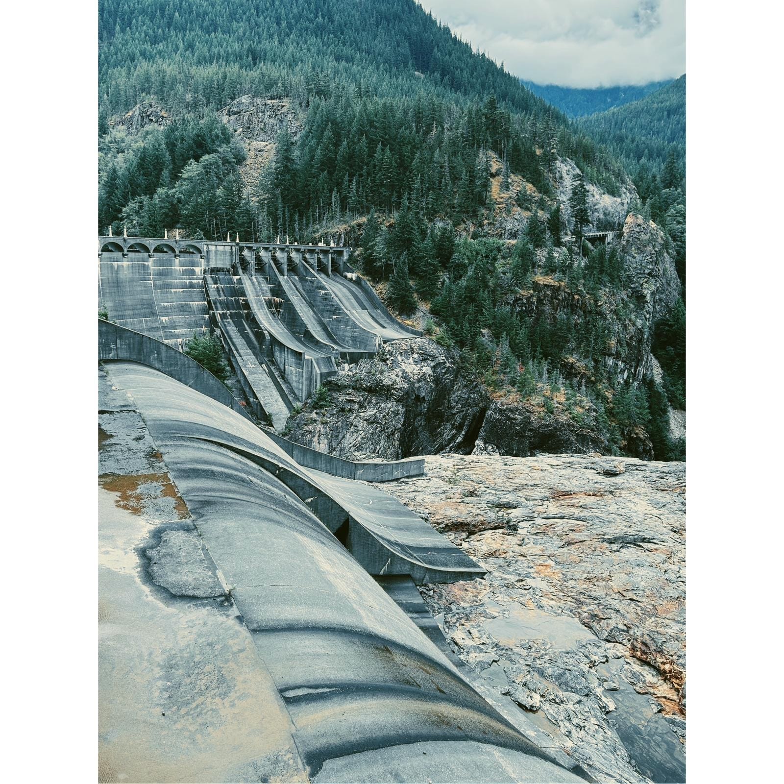 Concrete dam spillway with curved channels and forested mountain backdrop
