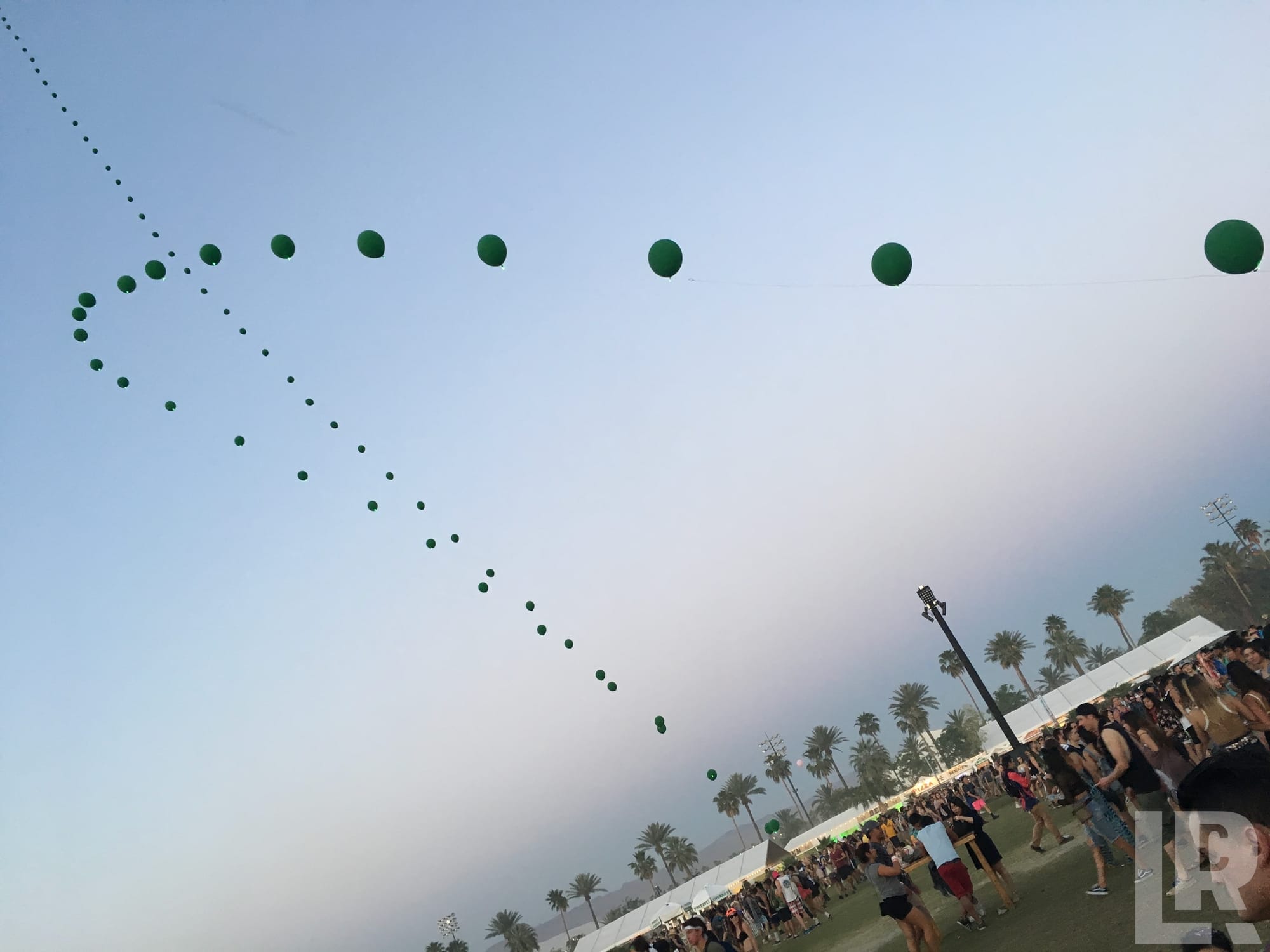 Green balloons strung across the sky above a crowded outdoor music festival with palm trees in the background