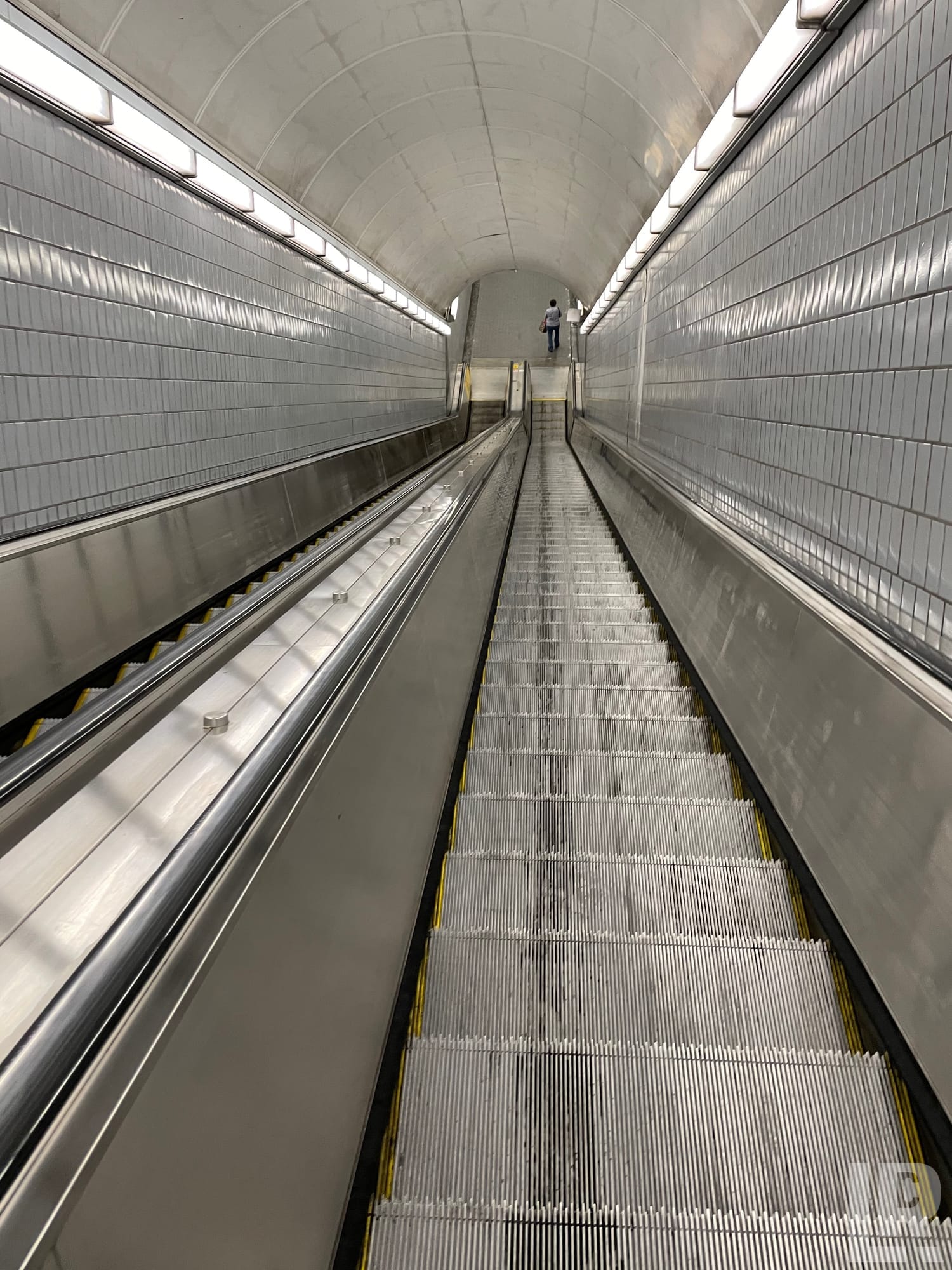 Upward view of a subway escalator inside a tiled tunnel with a person at the top