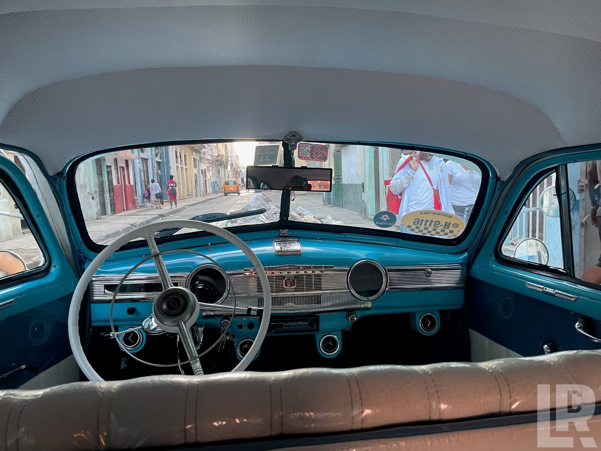 Rear passenger view inside a vintage teal Chevrolet with chrome dashboard and white steering wheel, looking out at a busy street