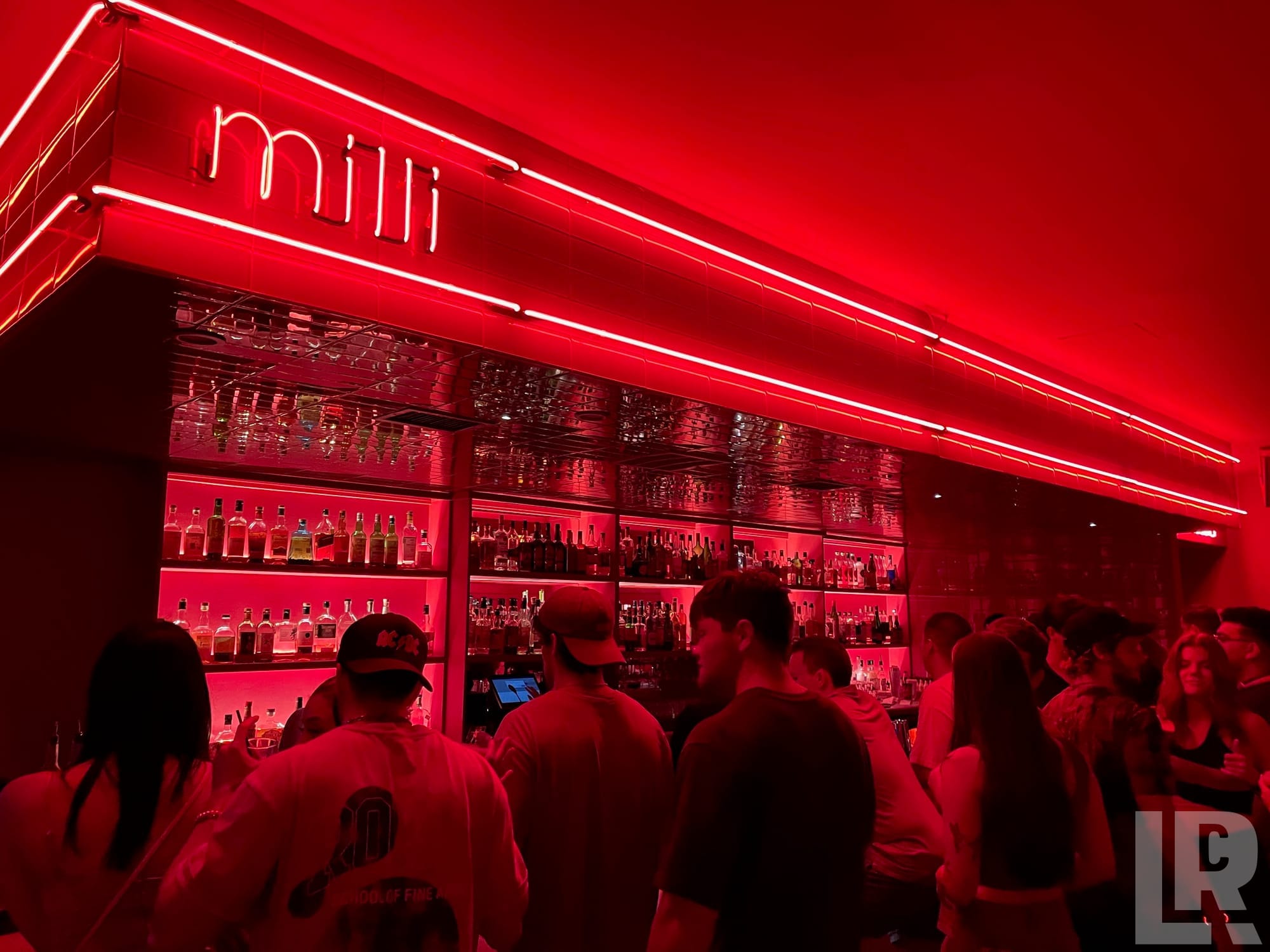 Red neon-lit bar interior named Milli with shelves of liquor and patrons gathered at the counter