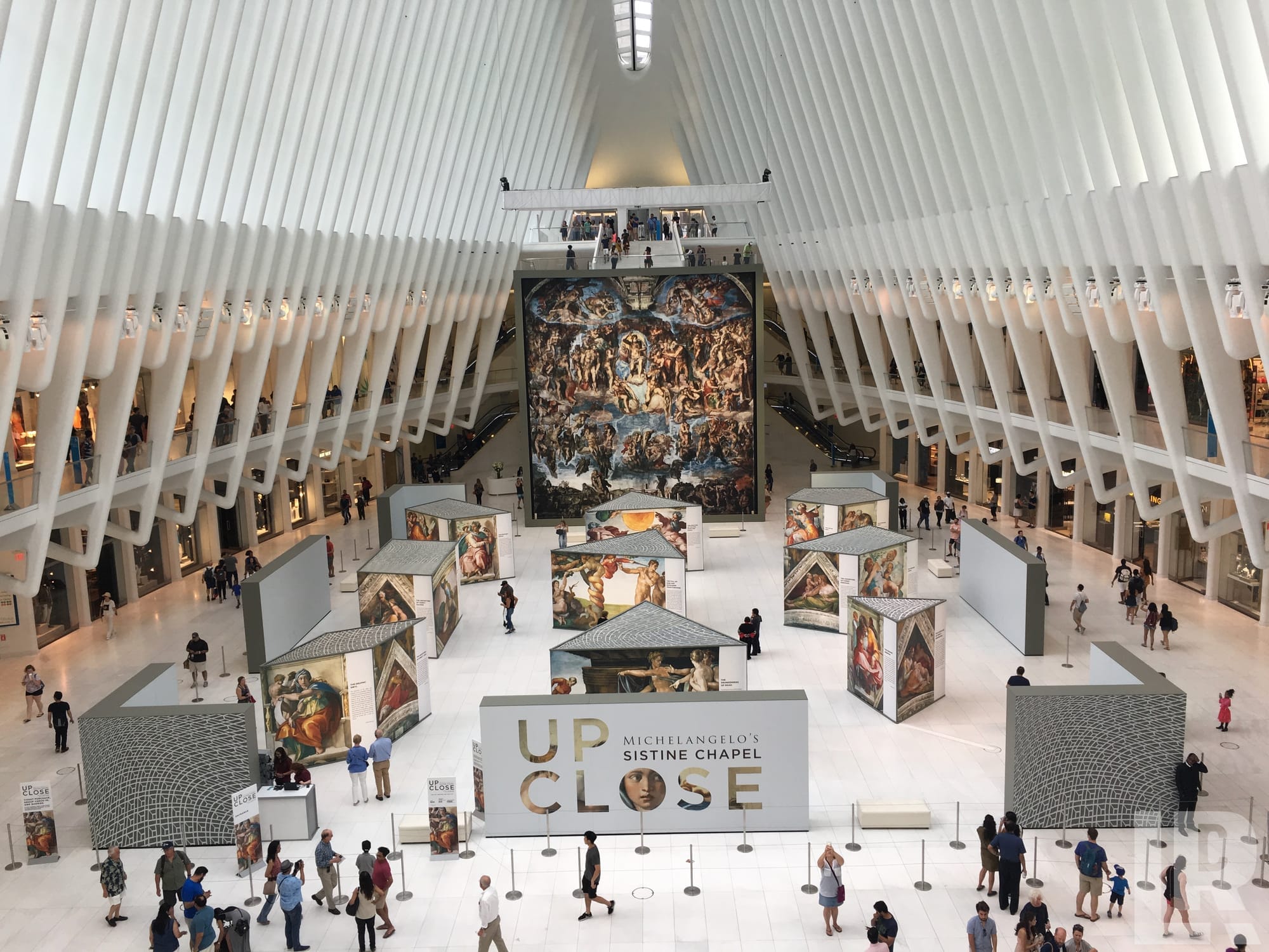 Interior of a large white ribbed architectural space displaying large reproductions of Michelangelo's Sistine Chapel frescoes with crowds of visitors