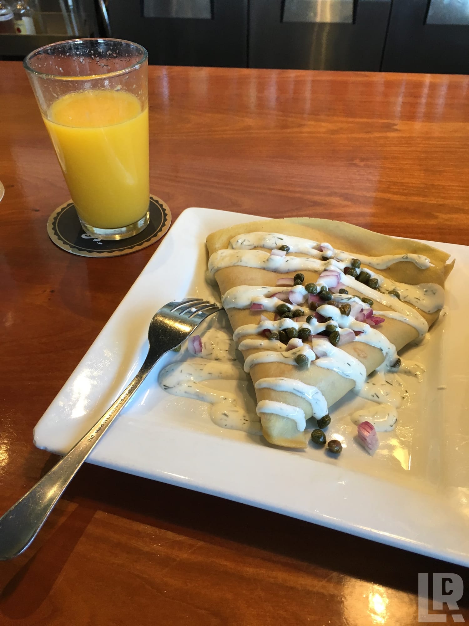 Savory crepes with cream sauce, capers, and red onion on a white plate next to a glass of orange juice