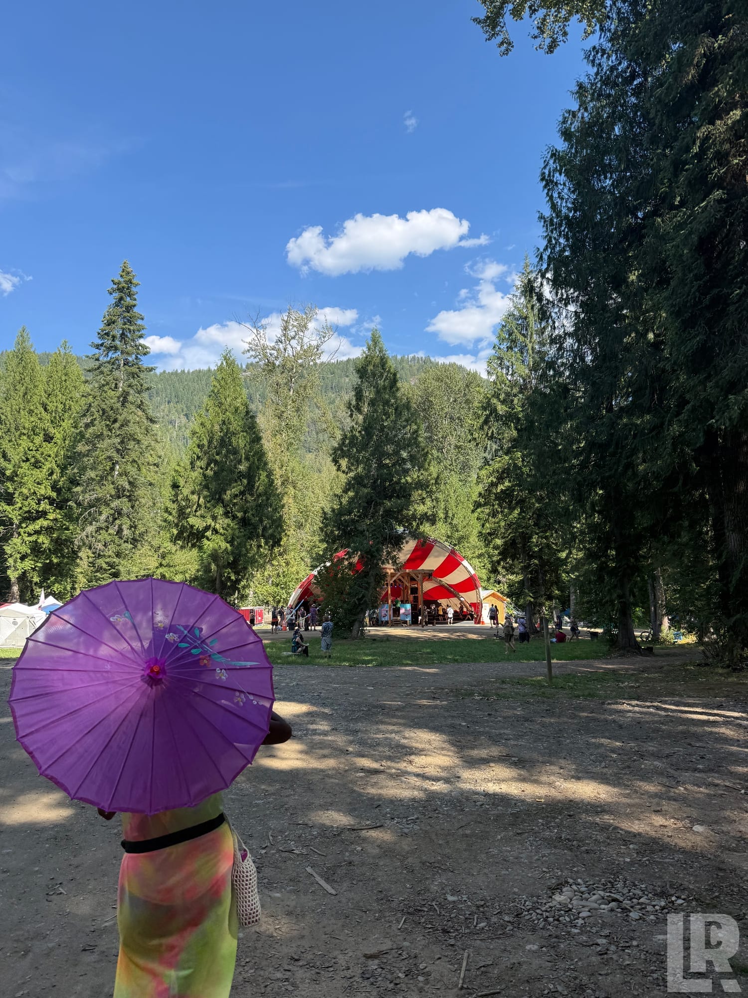 Person with purple parasol walking toward a striped festival tent in a forested mountain setting