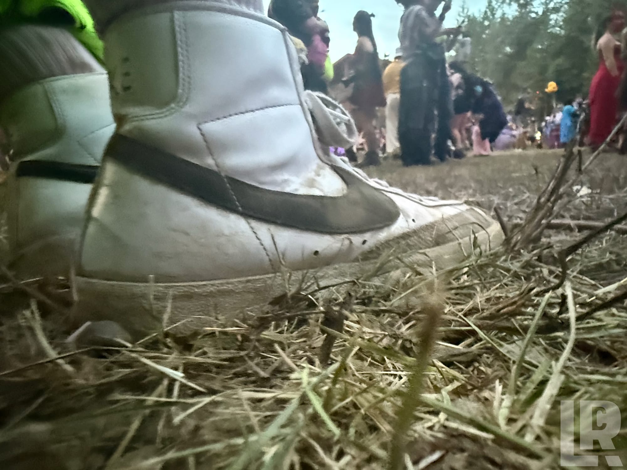 Close-up ground-level view of a white Nike high-top sneaker on dry grass with blurred crowd behind