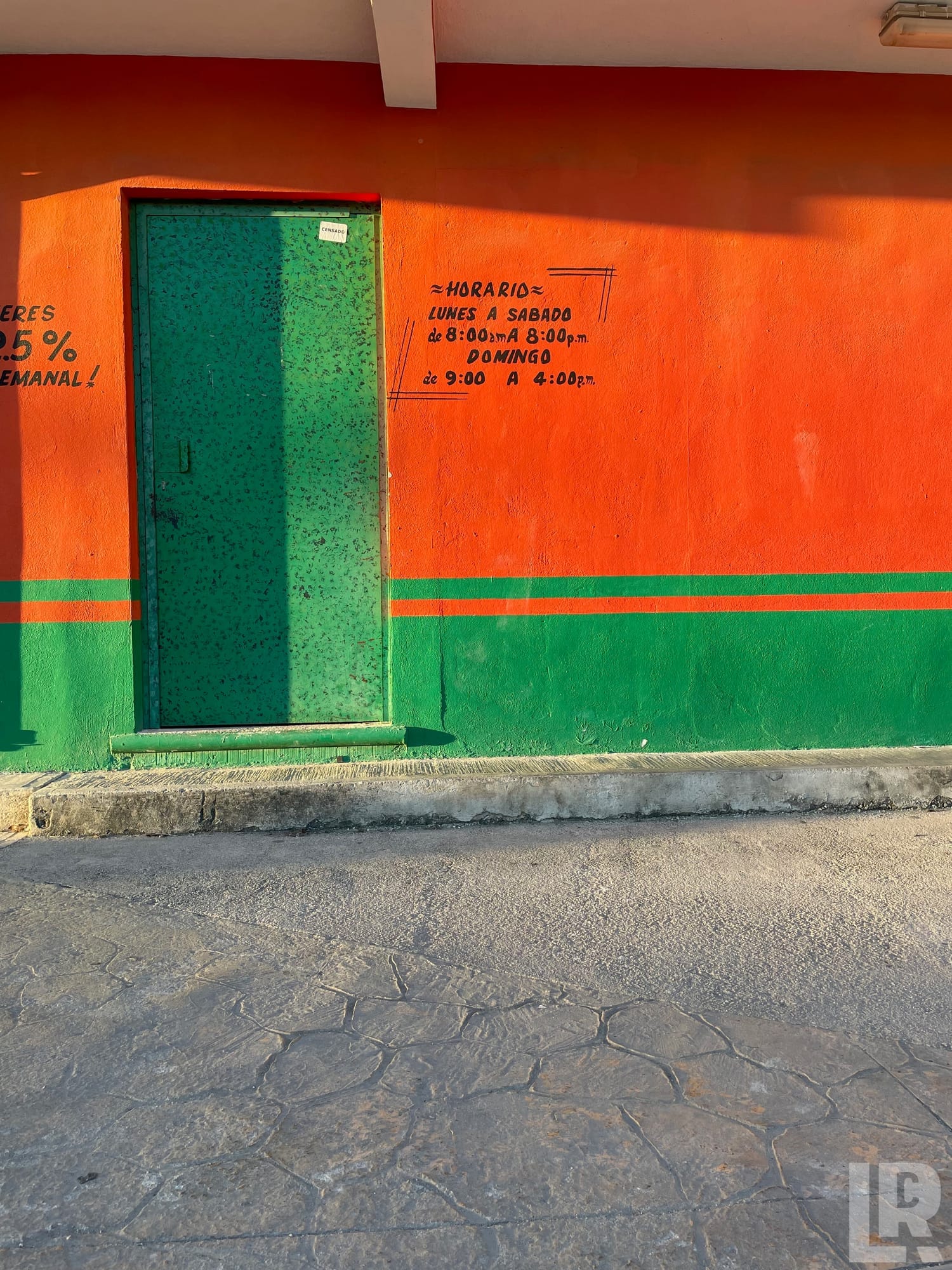 Orange and green painted storefront wall with green metal door and Spanish business hours painted on the wall