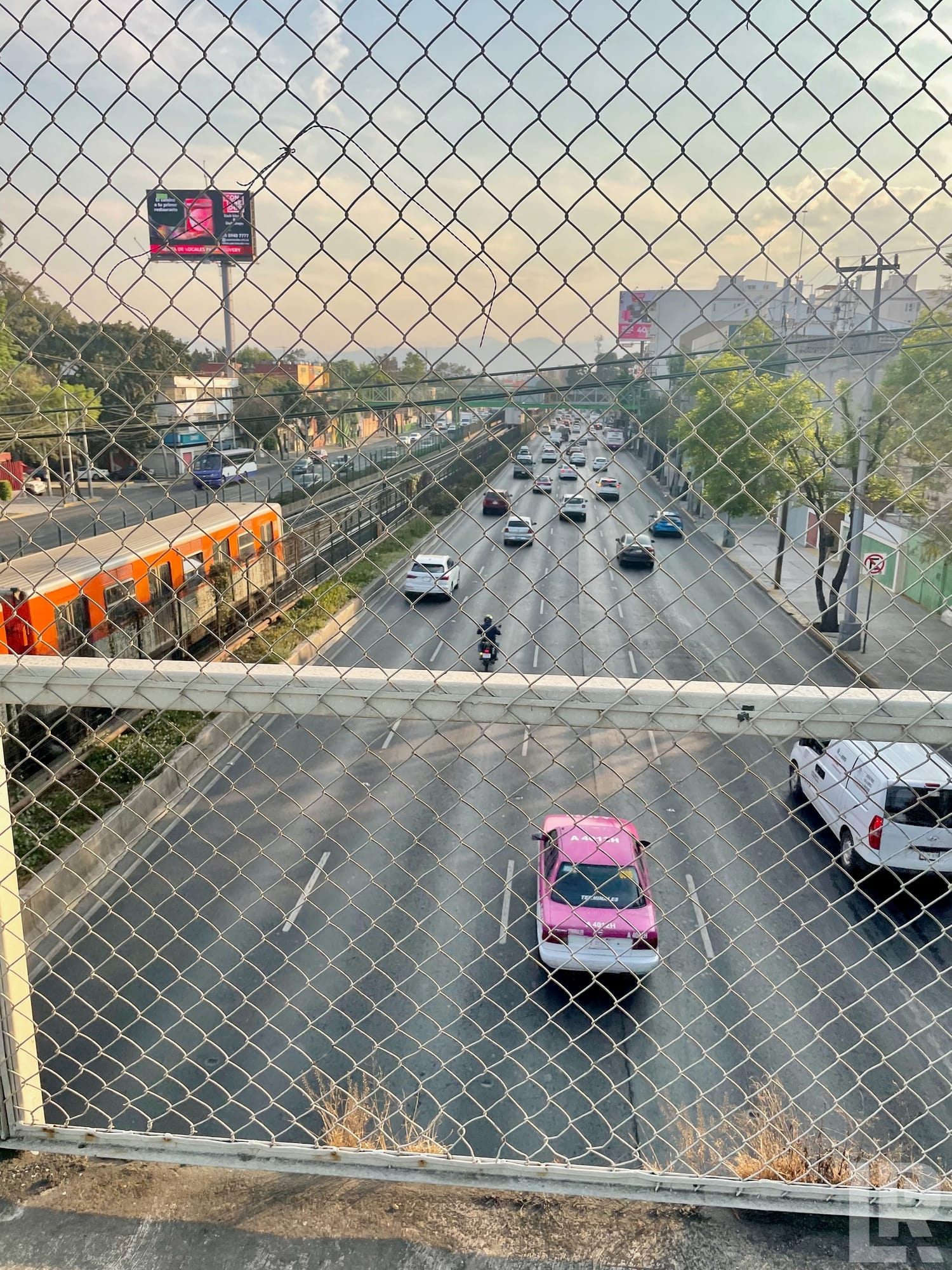Chain-link fence on an overpass overlooking a multi-lane urban highway with cars and an orange metro train