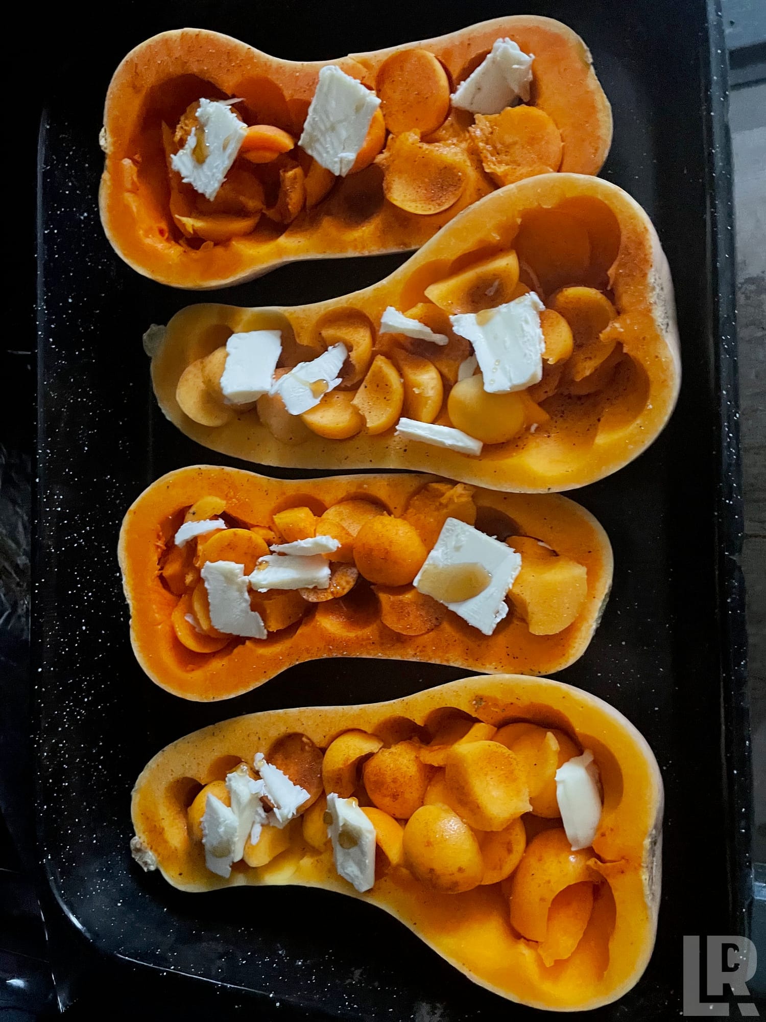 Four halved butternut squashes topped with carrot slices and cream cheese on a dark roasting pan