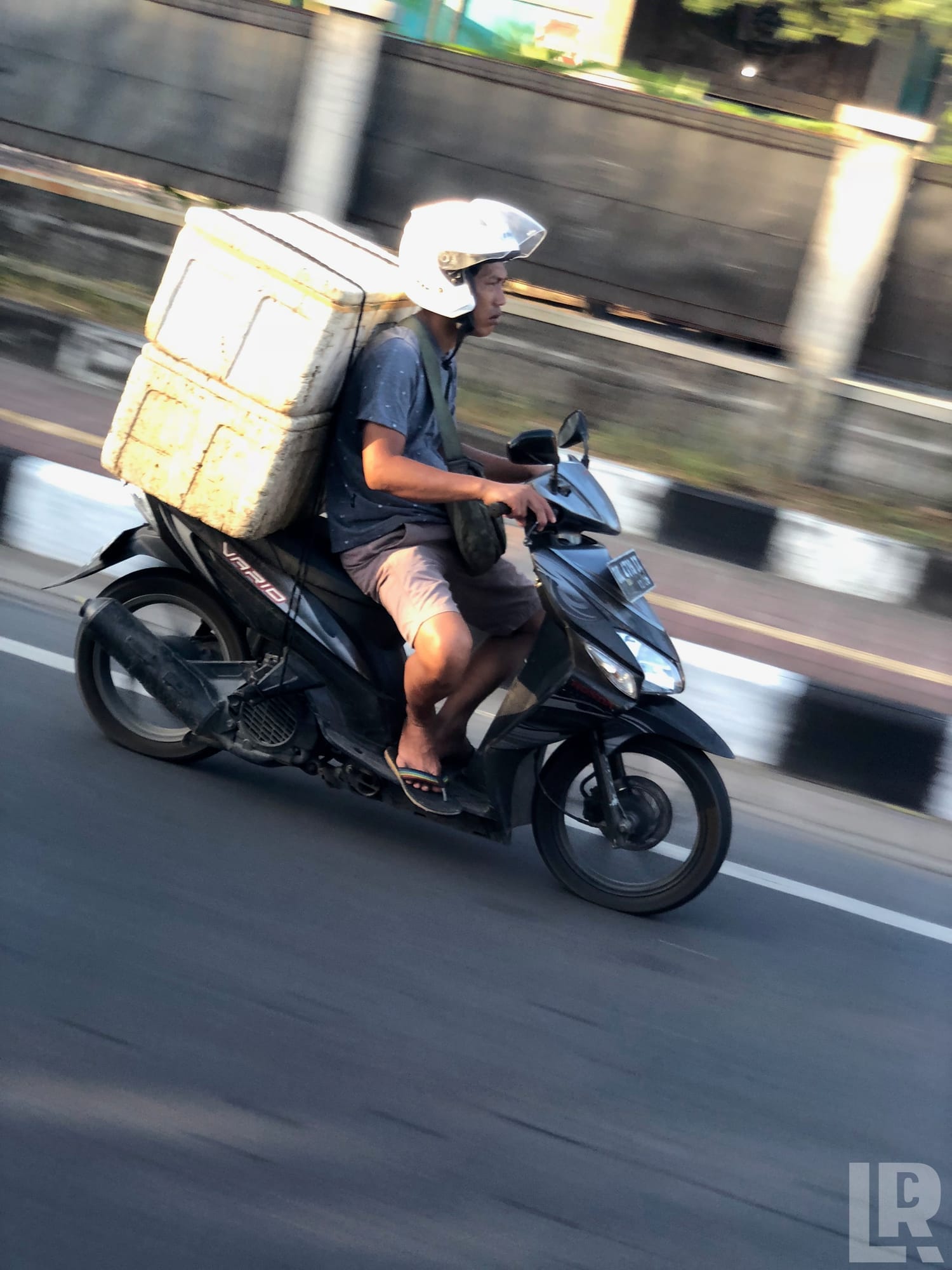 Man on black scooter carrying large styrofoam box on his back while riding on a road