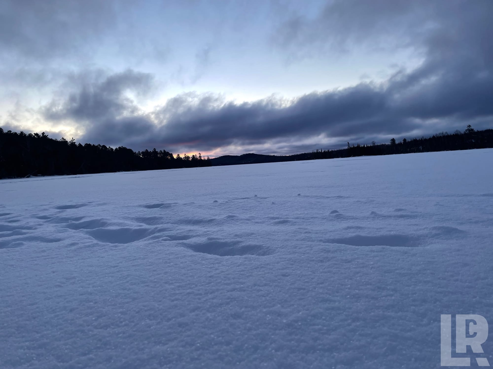 Snow-covered frozen lake at dusk with dark clouds and a forested shoreline in the background