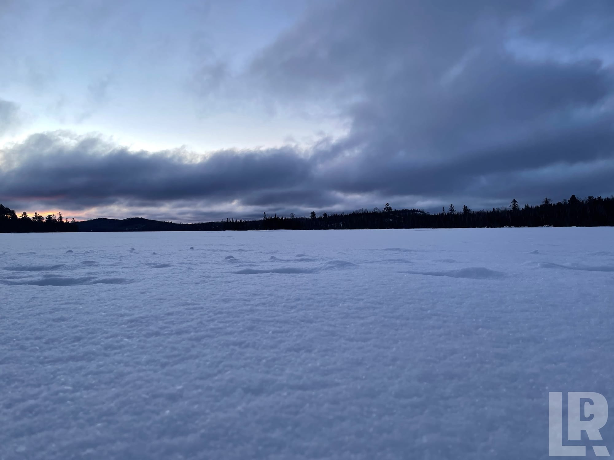 Snow-covered frozen lake at dusk with dark clouds and distant evergreen treeline