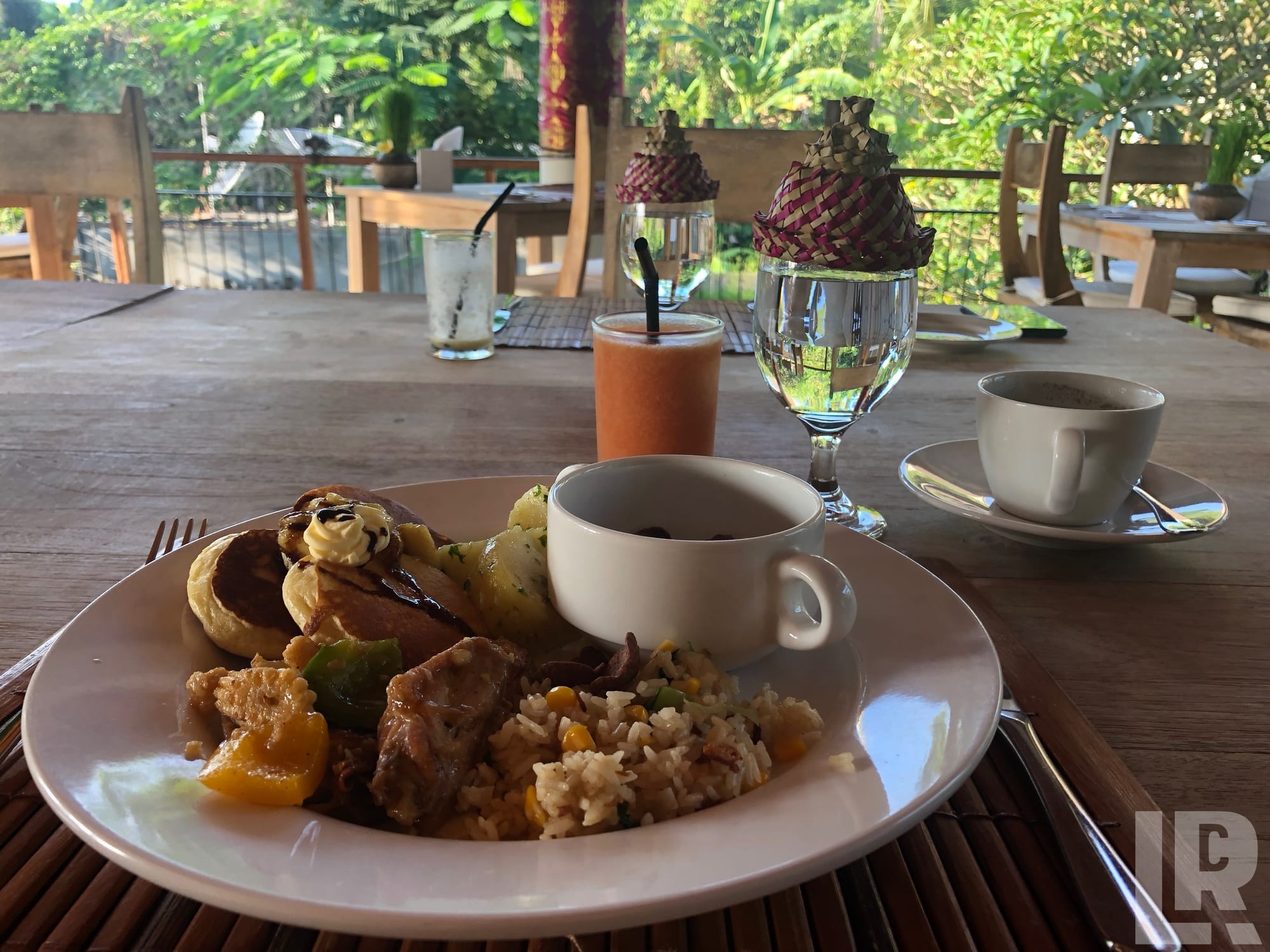 Plate of rice, meat, vegetables, and pancakes with coffee and juice drinks on a wooden table overlooking lush greenery