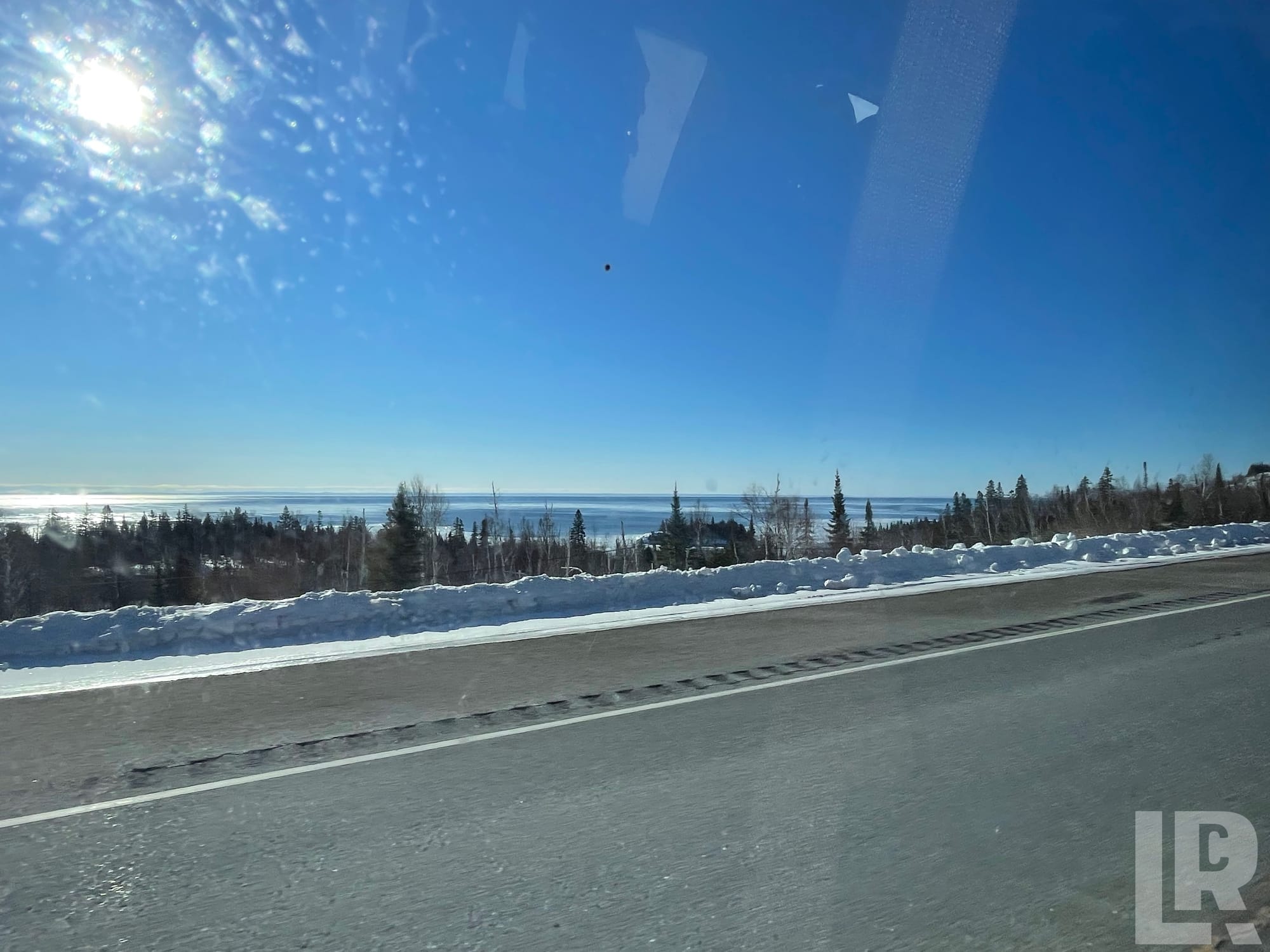Snow-lined road with conifer trees and frozen water body visible on a sunny winter day