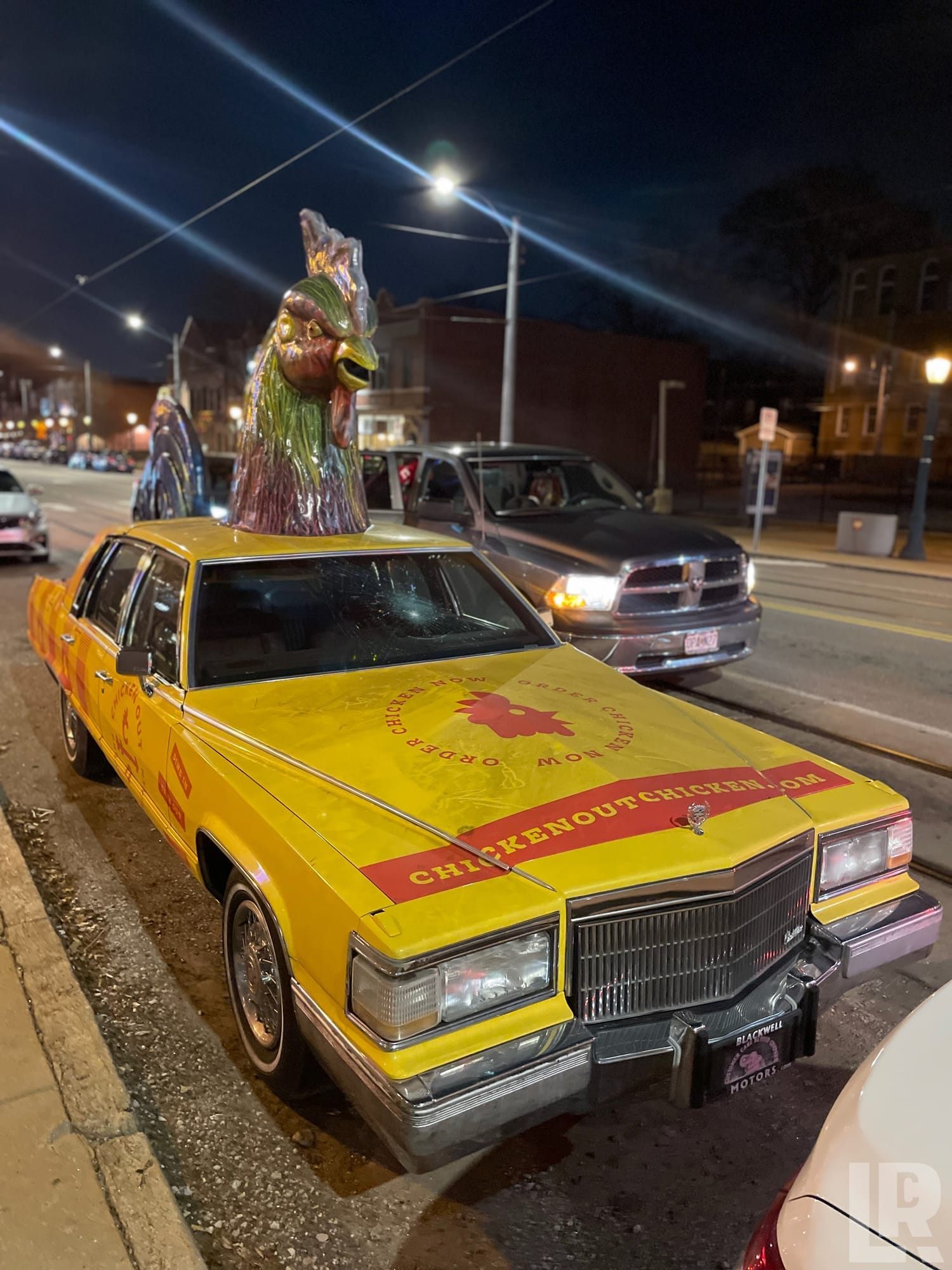 Yellow Cadillac car with giant chicken head sculptures on roof advertising Chicken Out, parked on urban street at night