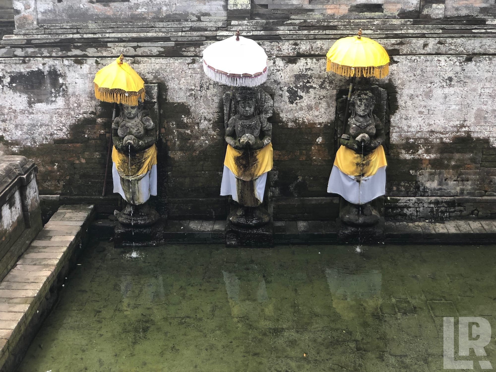 Three Balinese stone deity statues with yellow parasols and fabric draping, functioning as fountains over a green-tinted ritual pool
