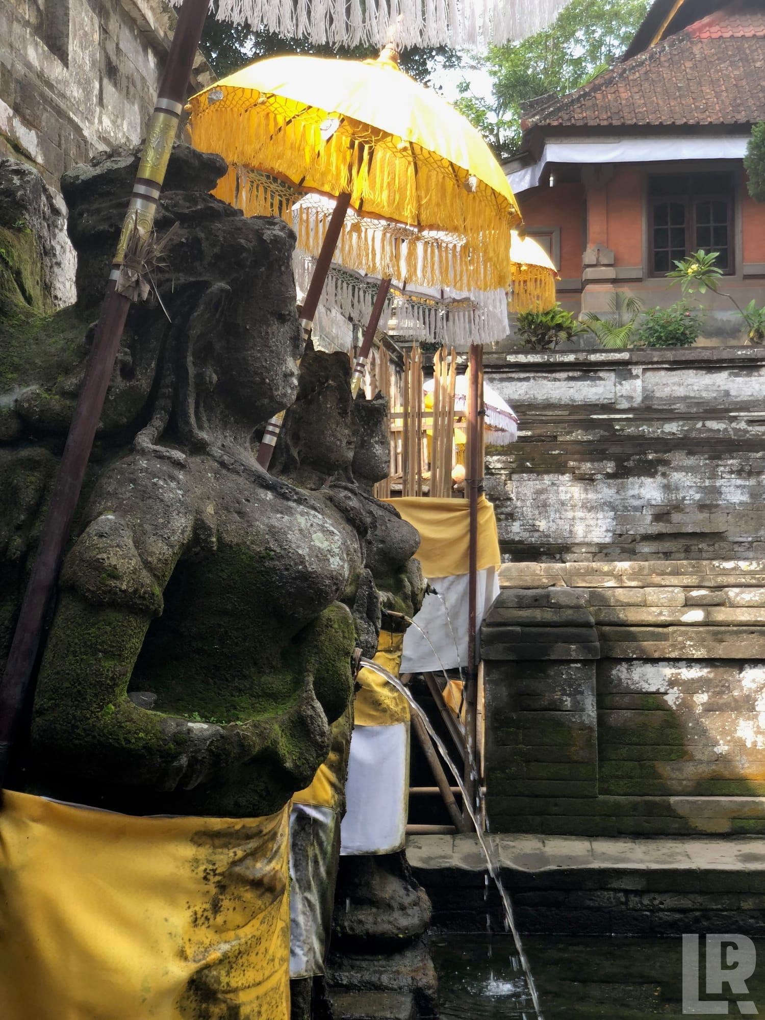 Moss-covered stone statue with yellow ceremonial cloth and golden fringed parasols at a Balinese Hindu temple