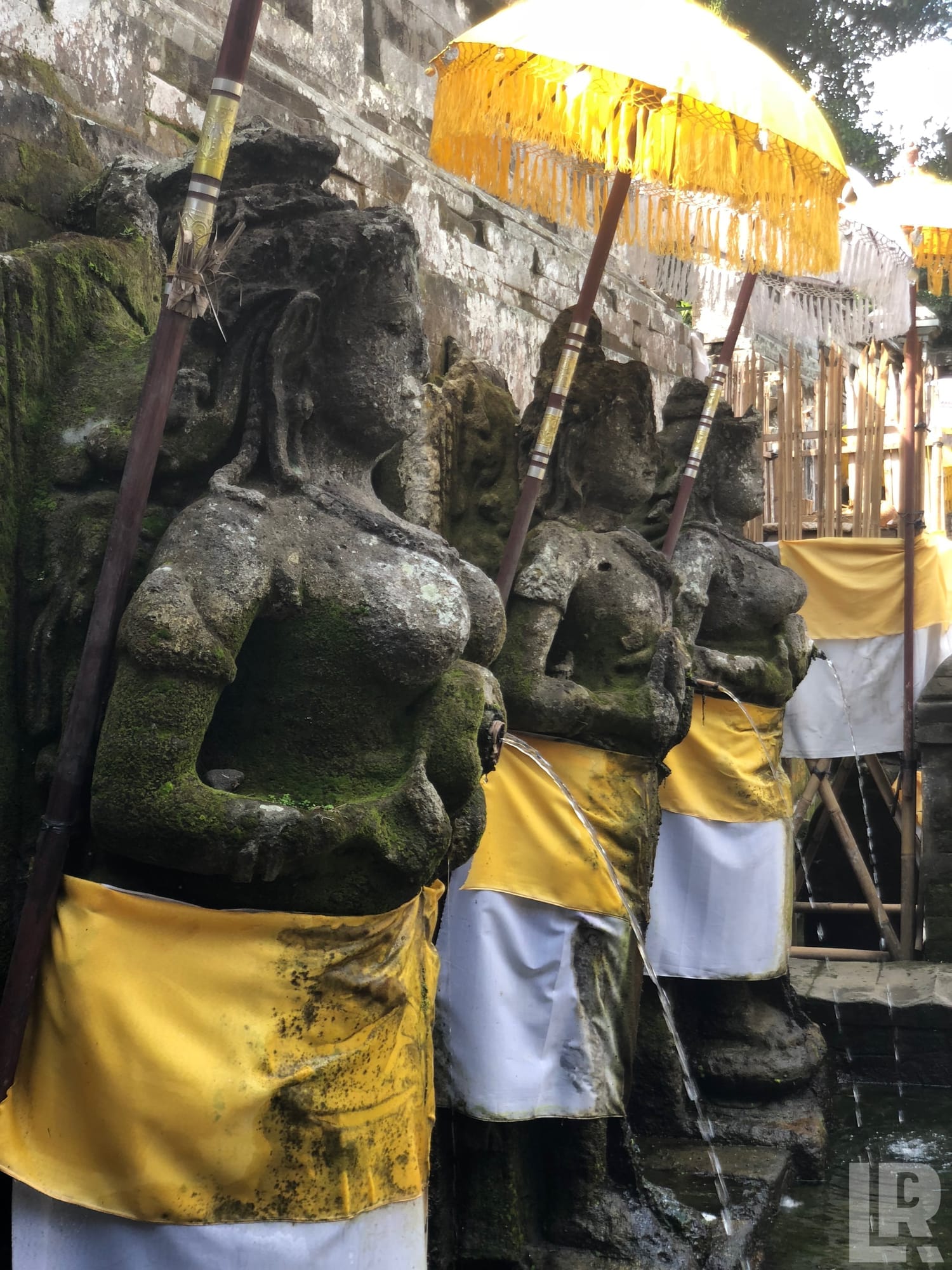 Moss-covered Balinese stone statues with yellow and white ceremonial cloths and water spouts at a temple