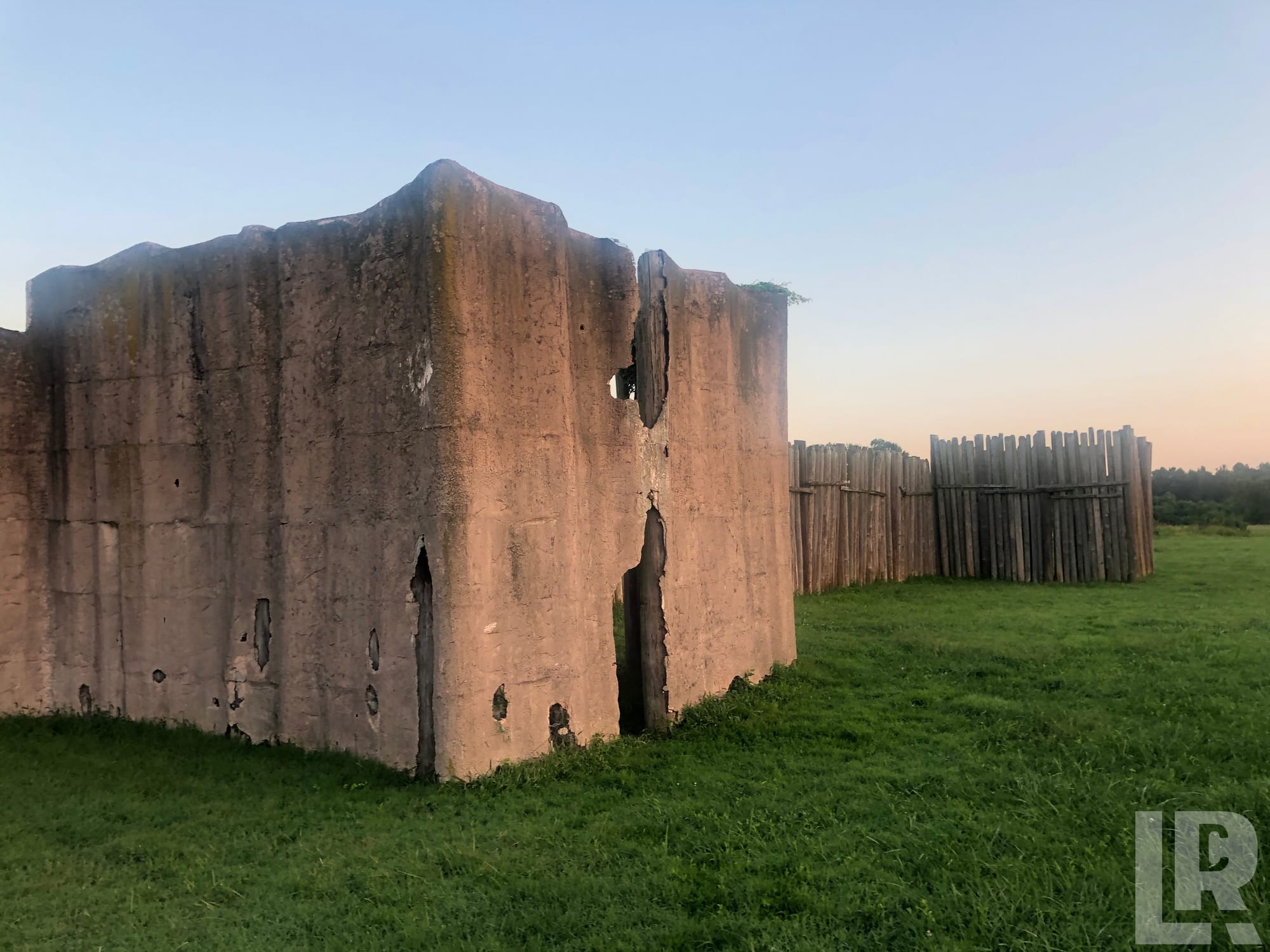 Deteriorating concrete wall ruins beside a wooden palisade fence on a grassy field