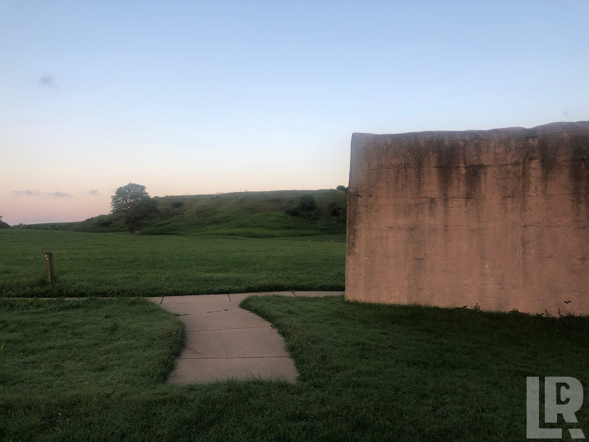Concrete slab beside a curved sidewalk with a grassy mound and trees in the background at twilight
