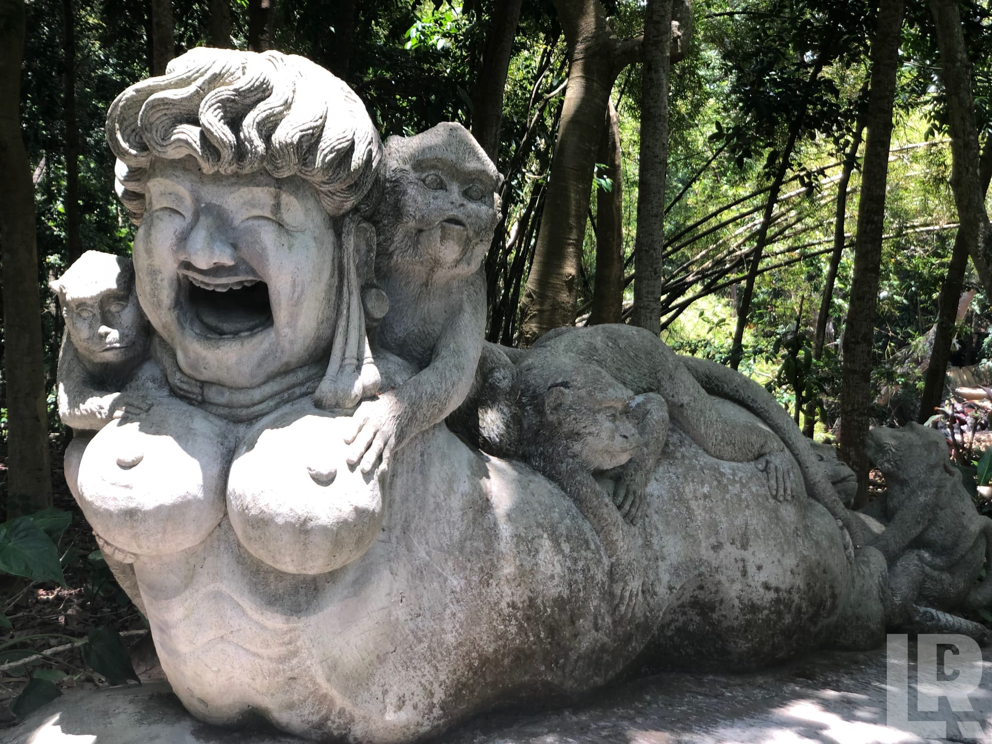 Large stone statue of a laughing reclining figure with monkeys carved around it in a forest