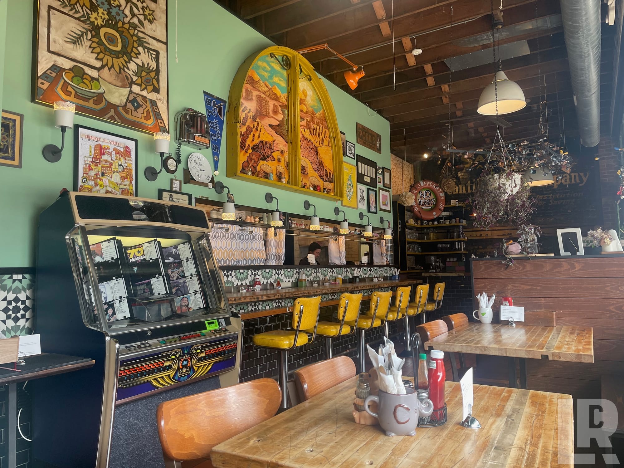 Retro diner interior with jukebox, yellow stools, green walls covered in artwork, and wooden tables