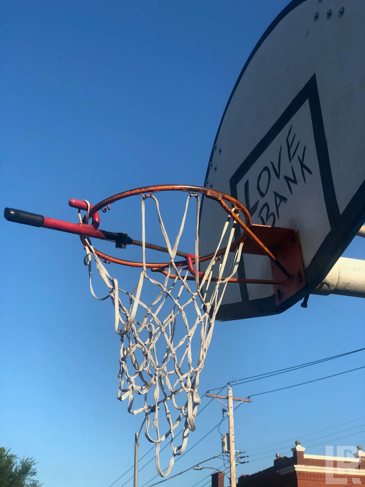 Close-up upward view of a rusty outdoor basketball hoop and net with 'LOVE BANK' graffiti on the backboard