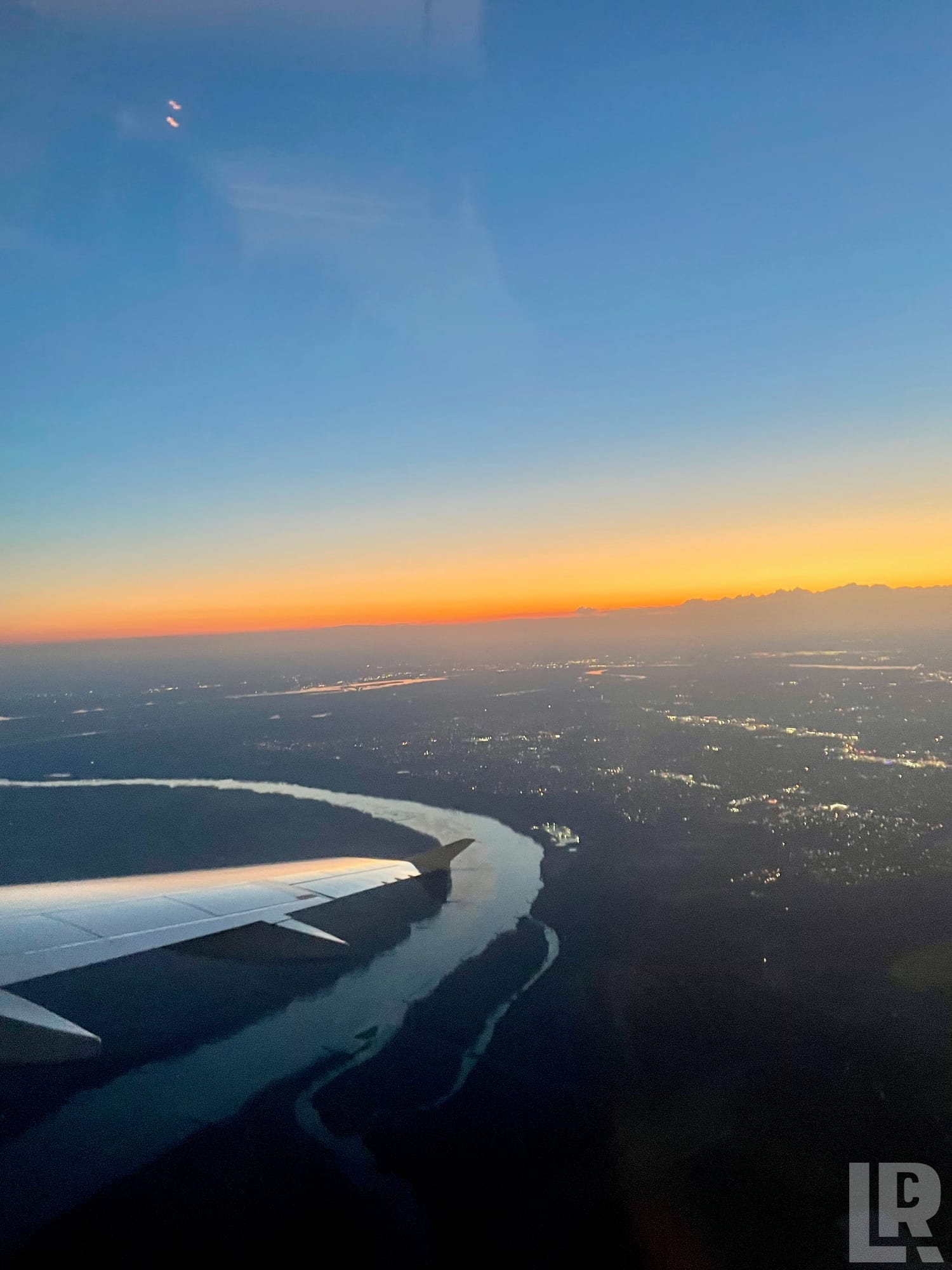 Airplane wing over a river and city at sunset with mountains in the distance