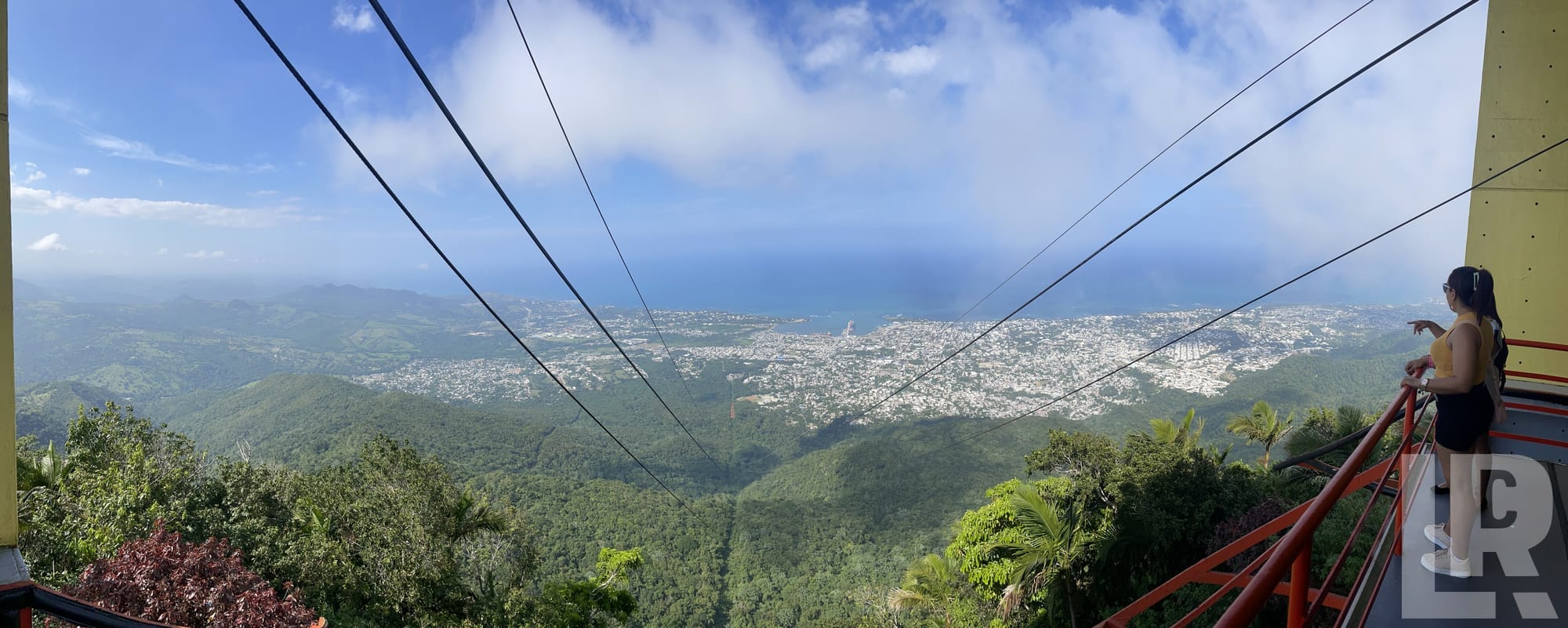 Woman on mountain observation platform overlooking tropical coastal city with cable car wires in foreground