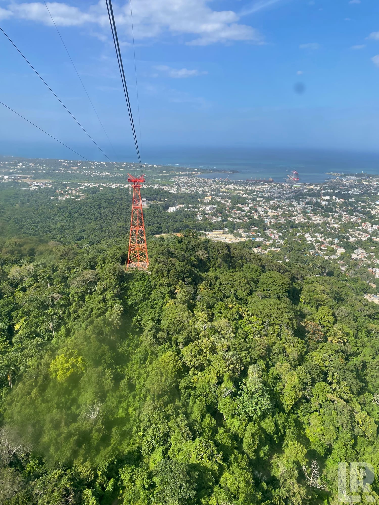 View from a cable car gondola over a forested hillside with a red support tower and a coastal city below