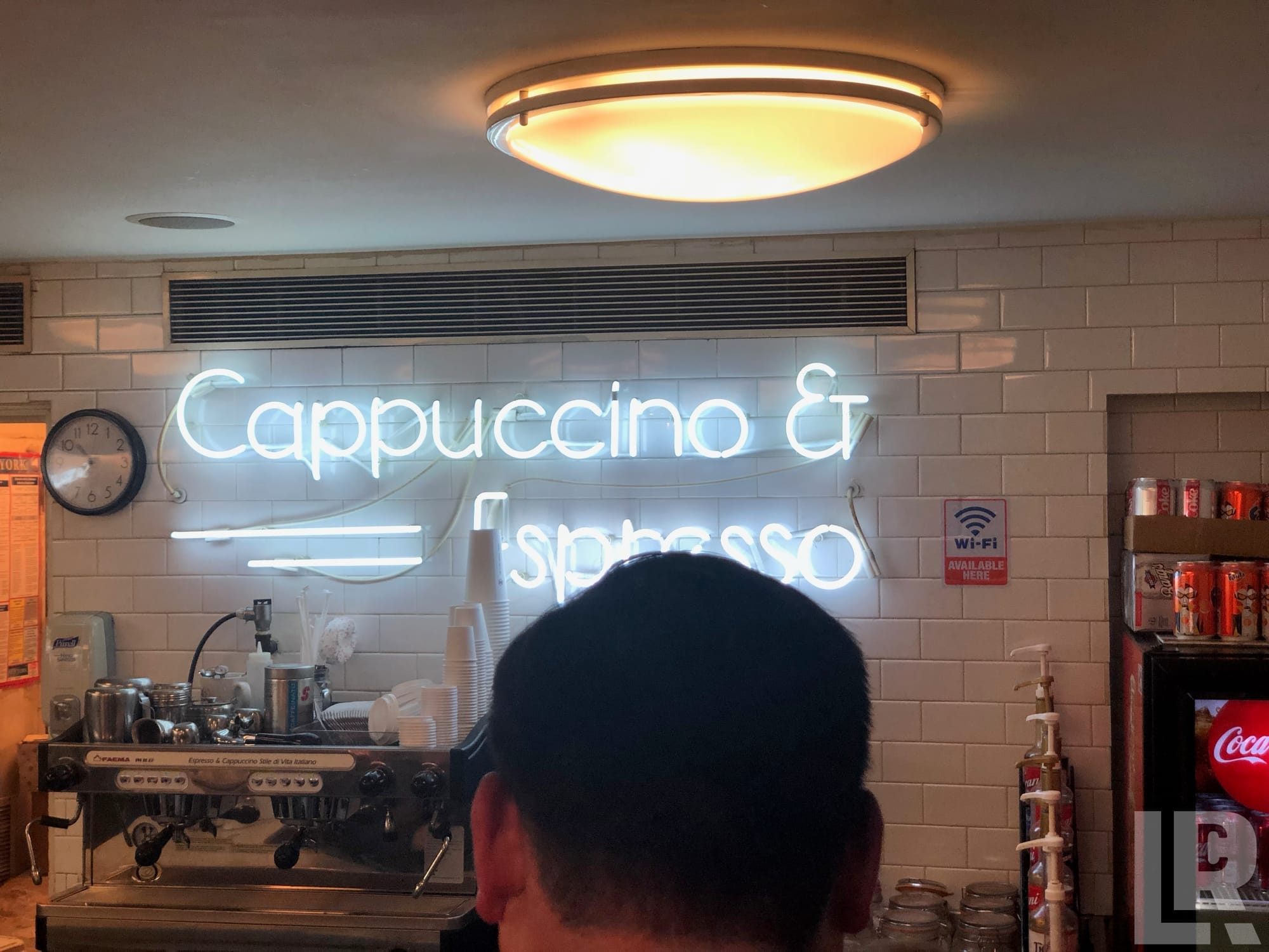 White neon cappuccino and espresso sign on subway tile wall above a commercial espresso machine in a cafe