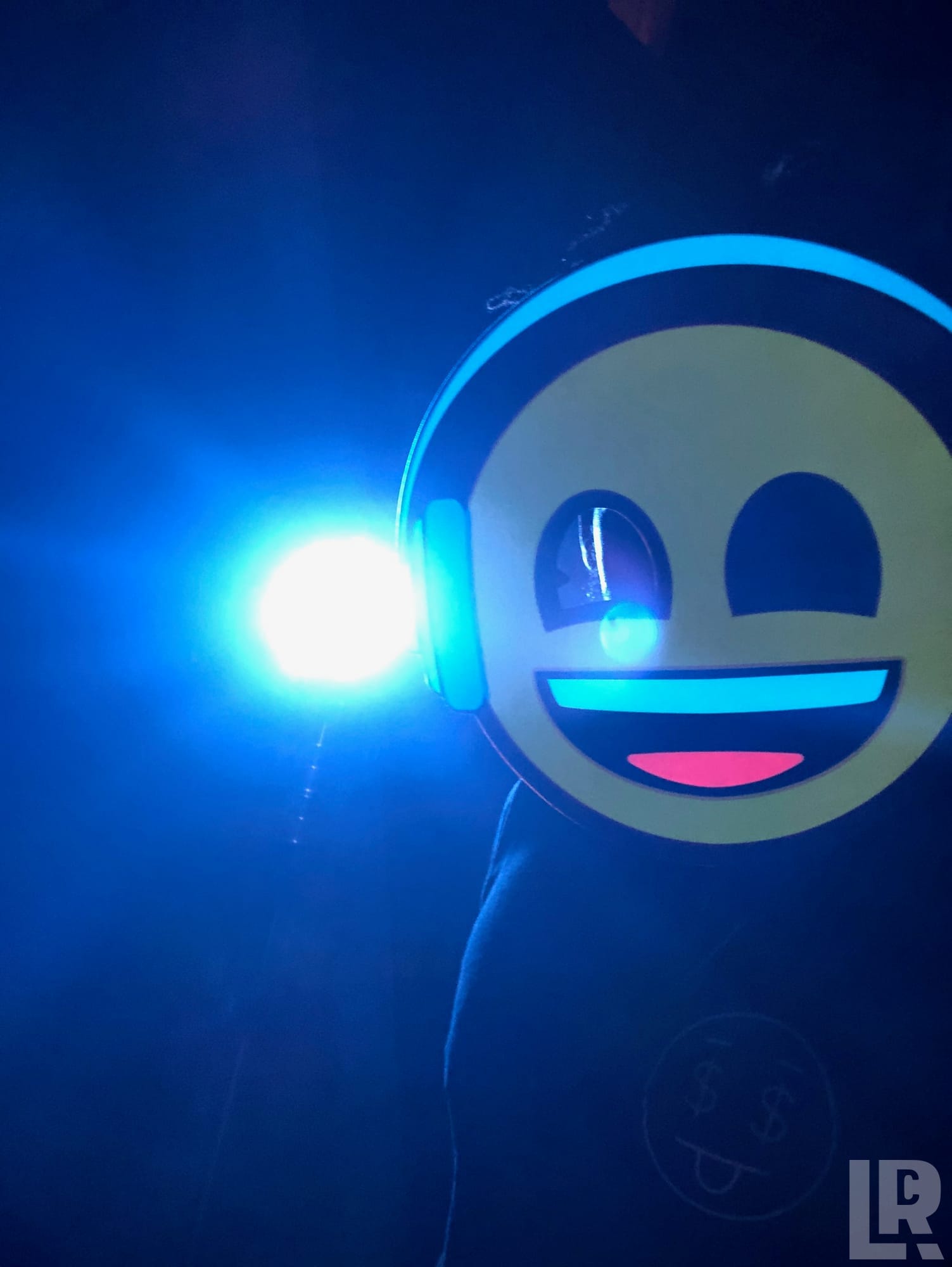 Person in illuminated emoji face mask with neon headphones under blue stage lighting