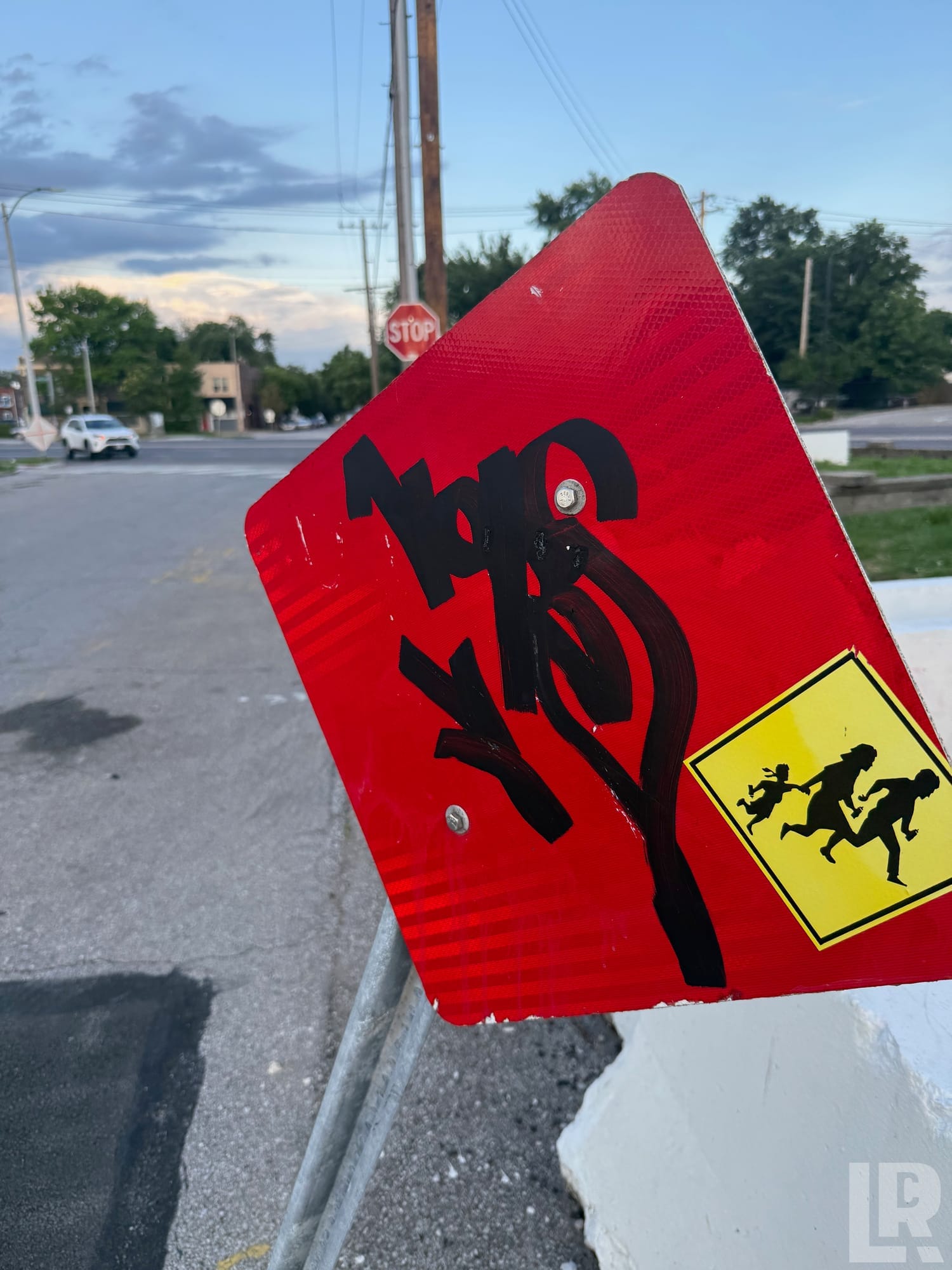 Red road sign with black graffiti and a yellow pedestrian crossing sticker at a street corner