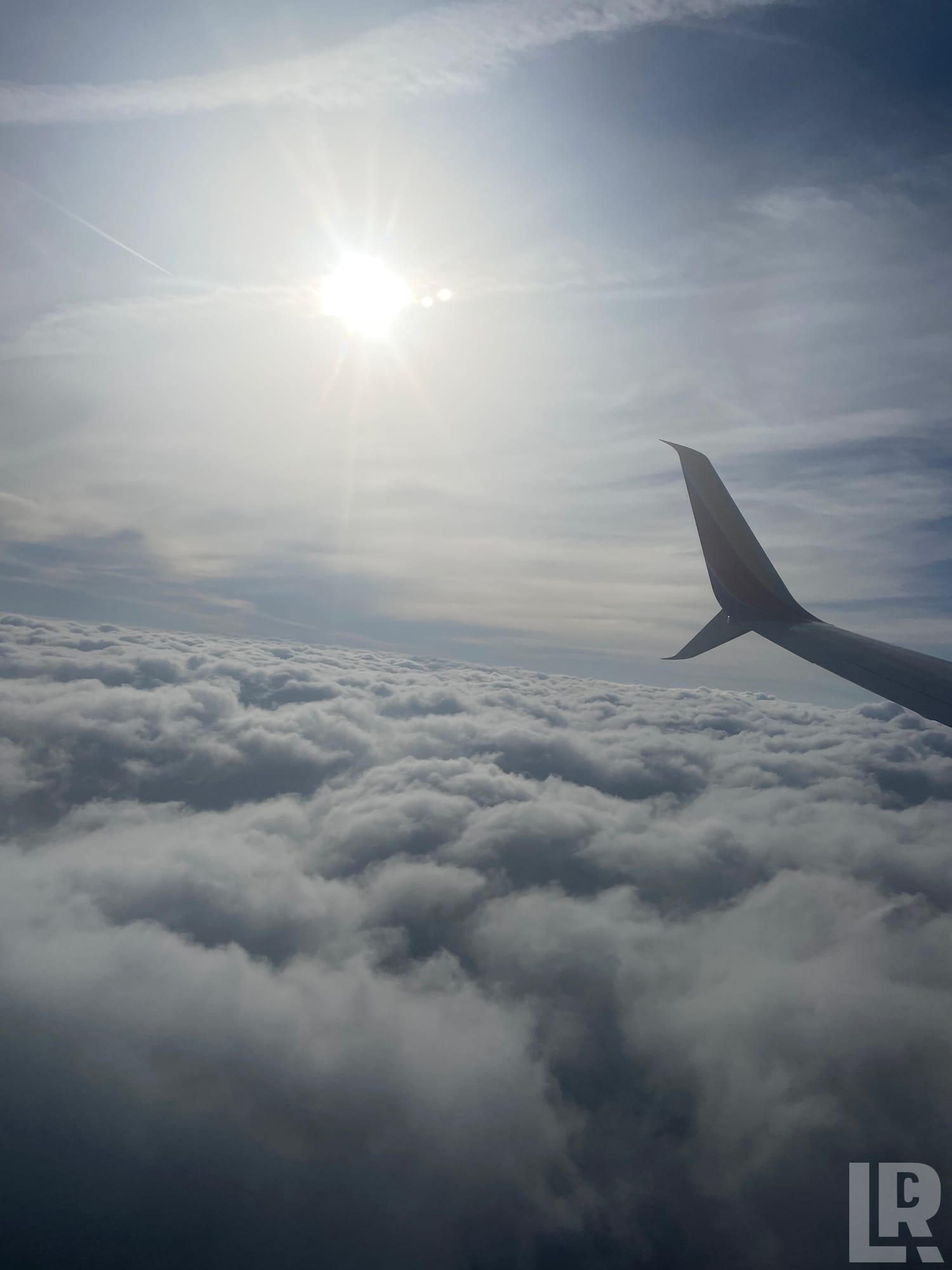 Airplane winglet above clouds seen from window seat with sun in blue sky