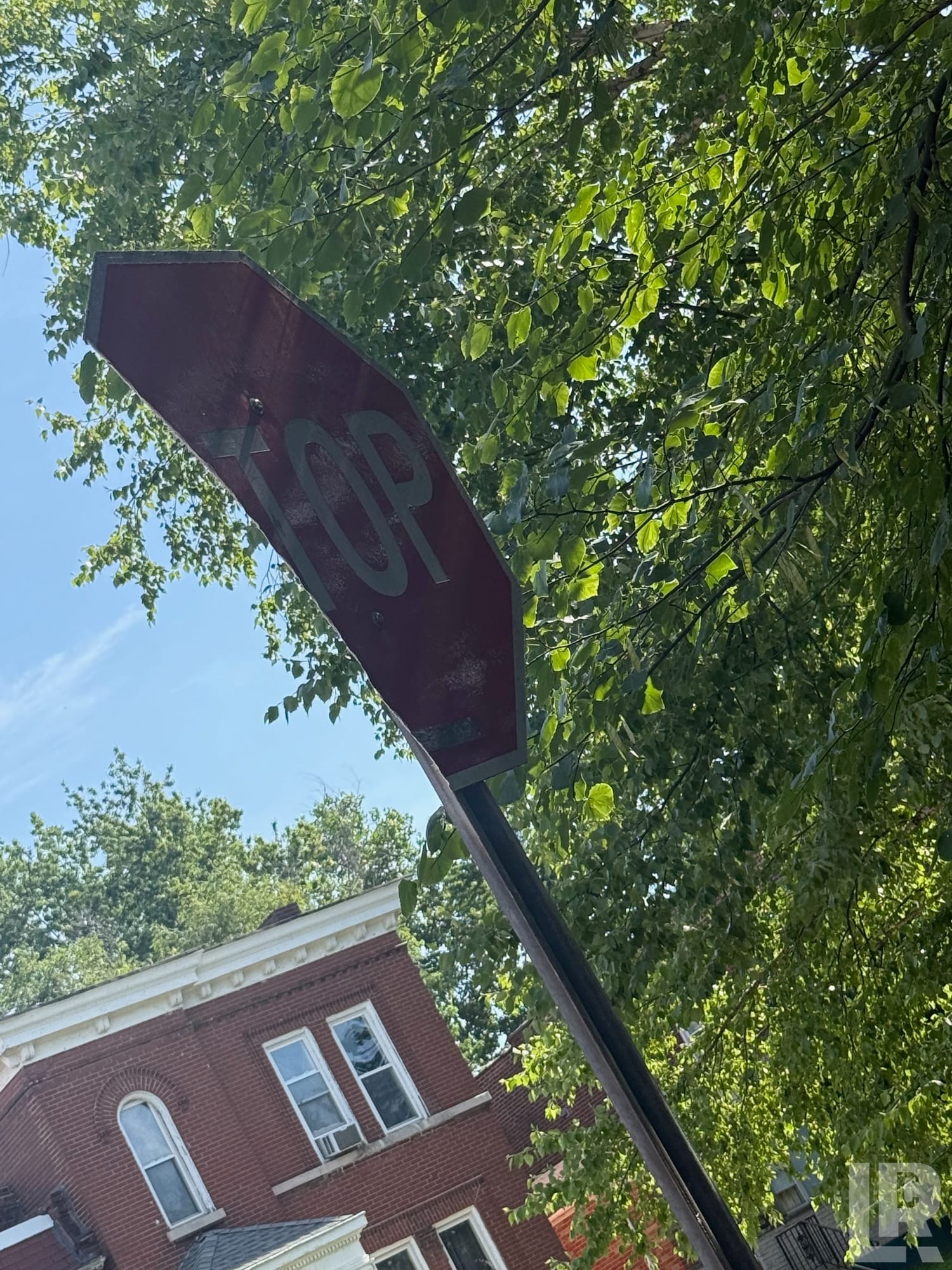 Faded red stop sign viewed from below against green tree canopy and brick building