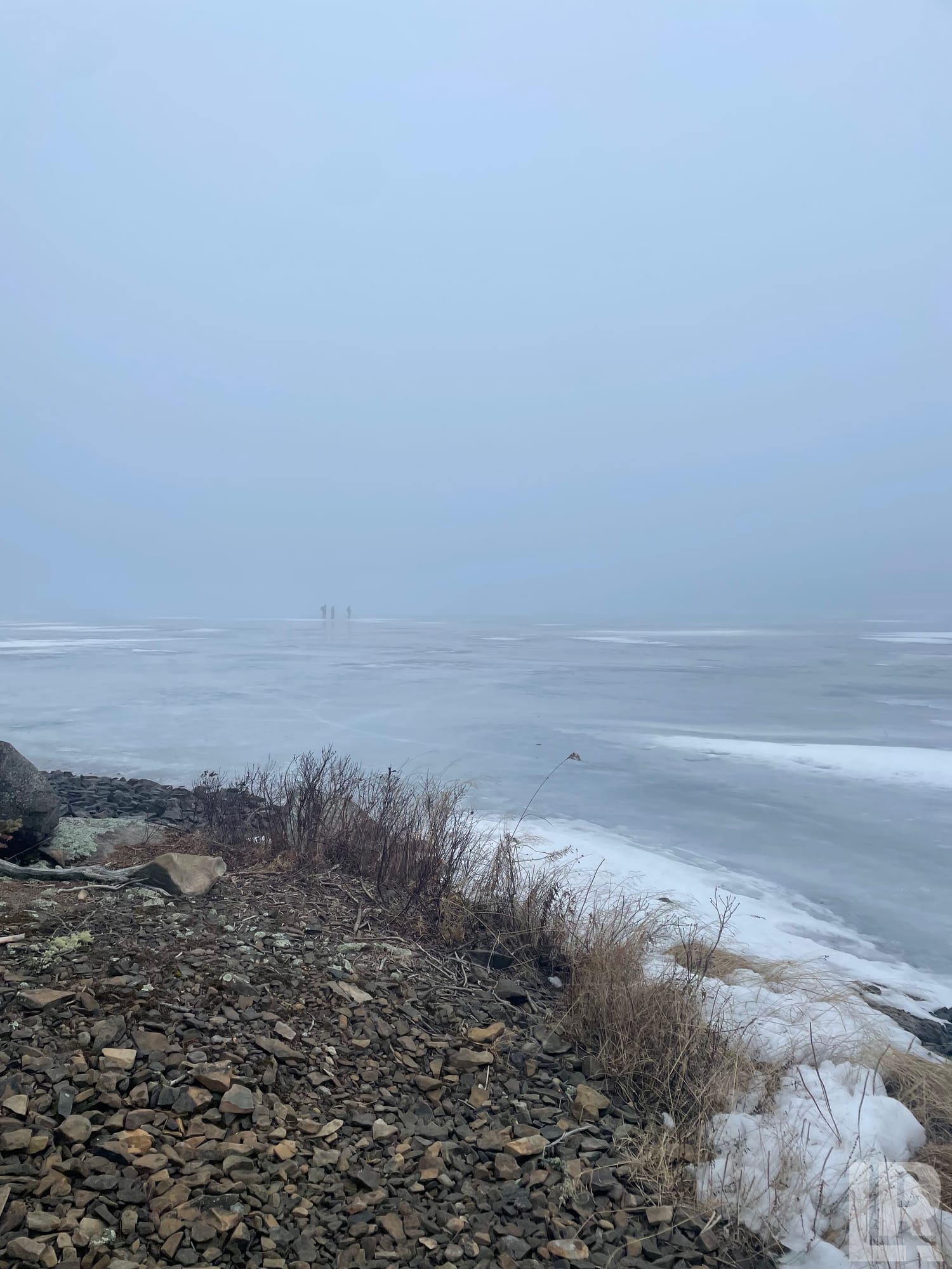 Foggy frozen lake with rocky shore, bare shrubs, snow patches, and two distant figures on the ice