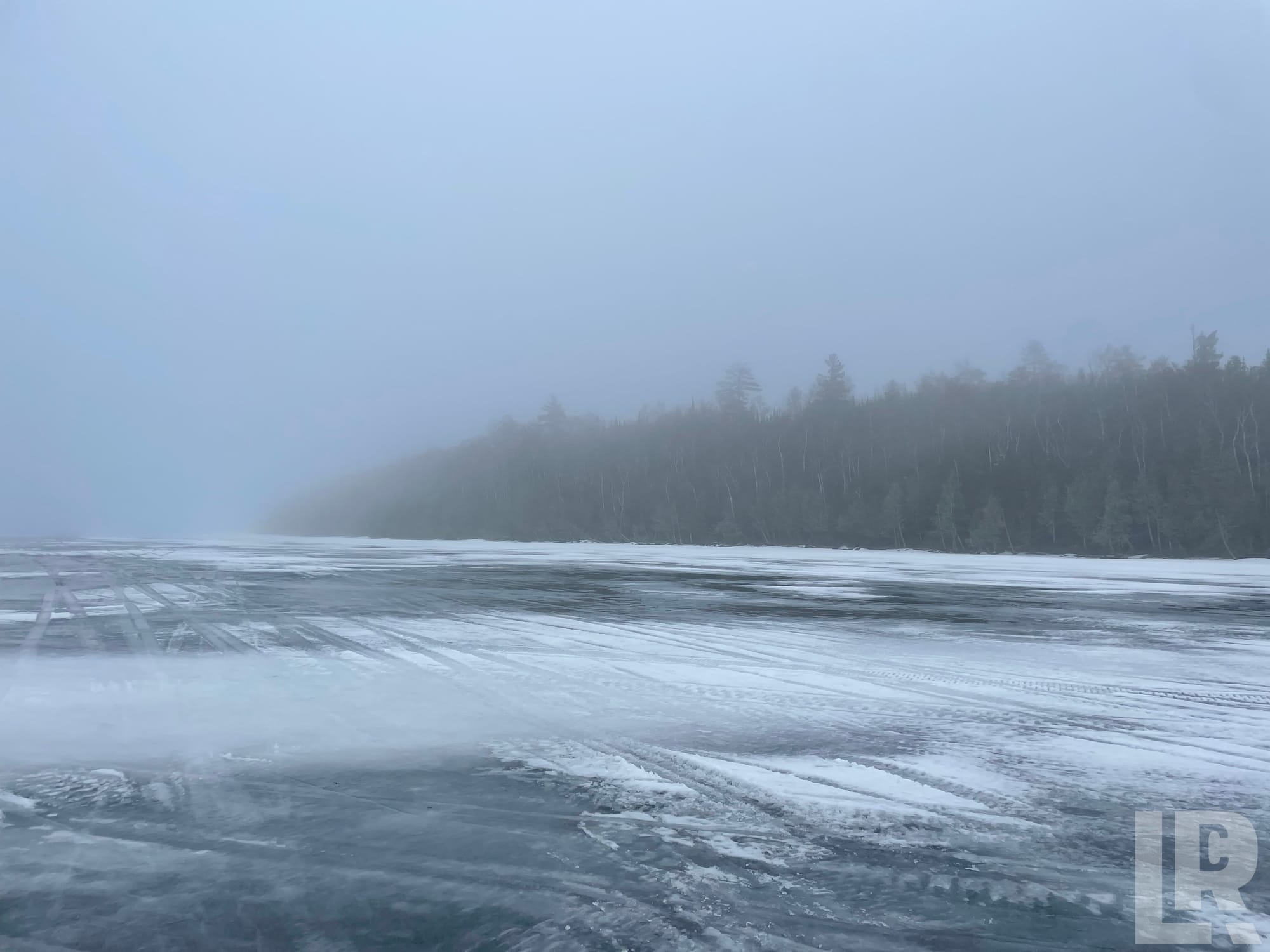 Icy frozen lake with snow drifts and tire tracks, misty forest in background