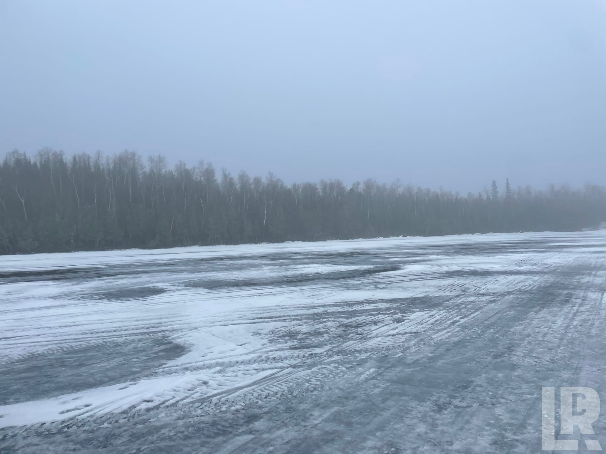 Icy frozen water surface with vehicle tracks leading toward a misty treeline in winter