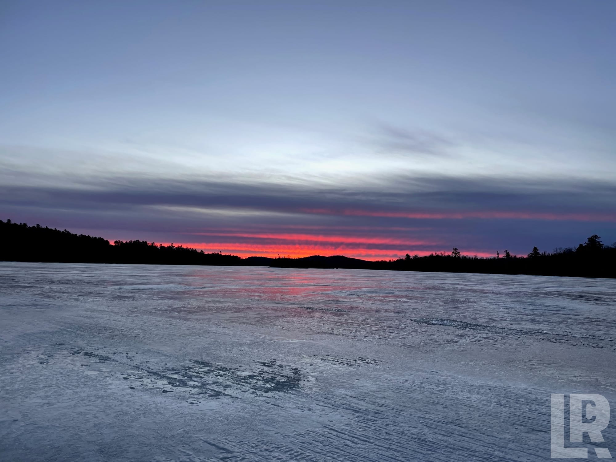 Frozen lake at sunset with bright red sky reflecting on the ice and silhouetted treeline