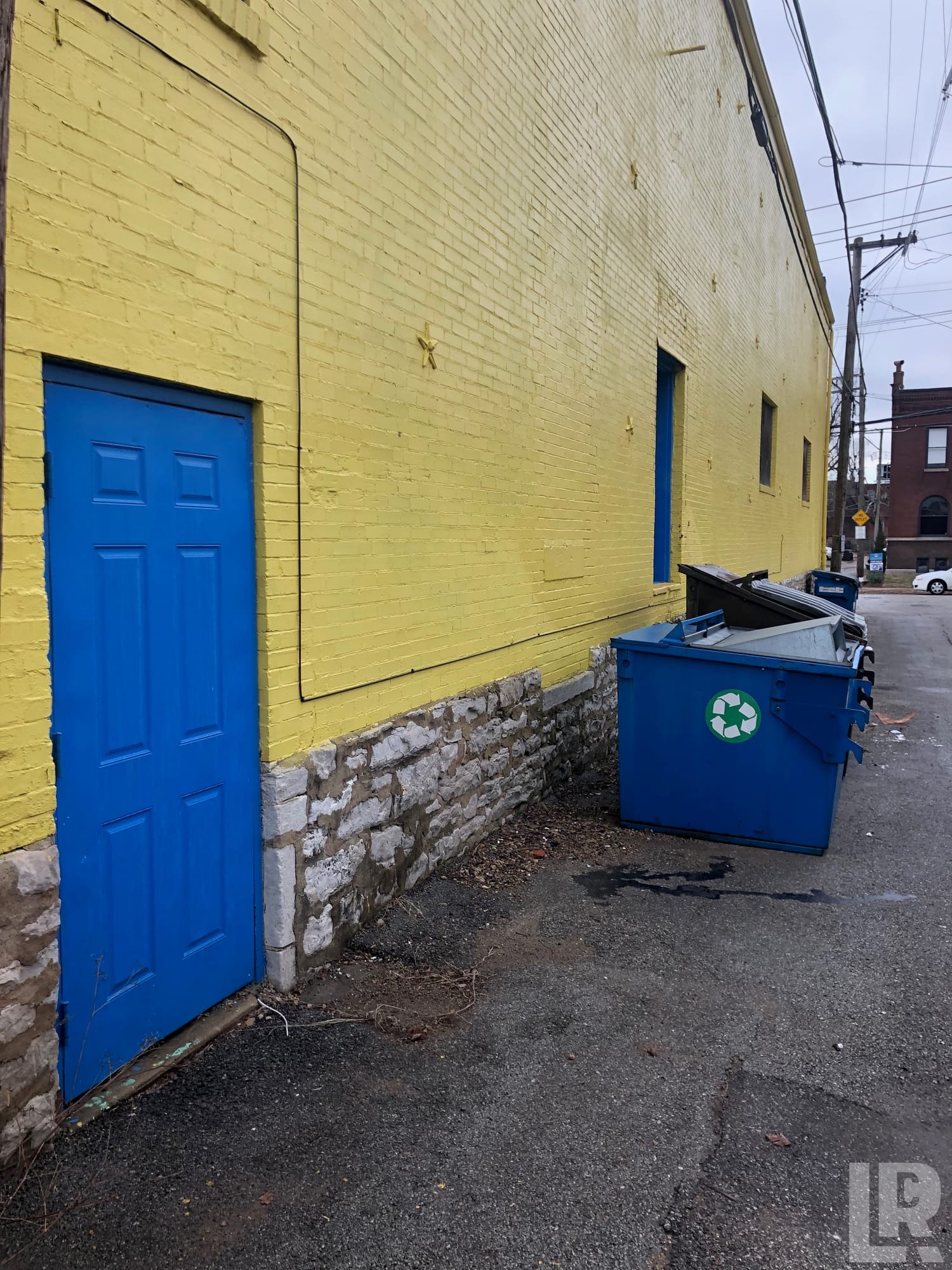 Yellow brick building exterior with blue door and blue recycling dumpster in an alley