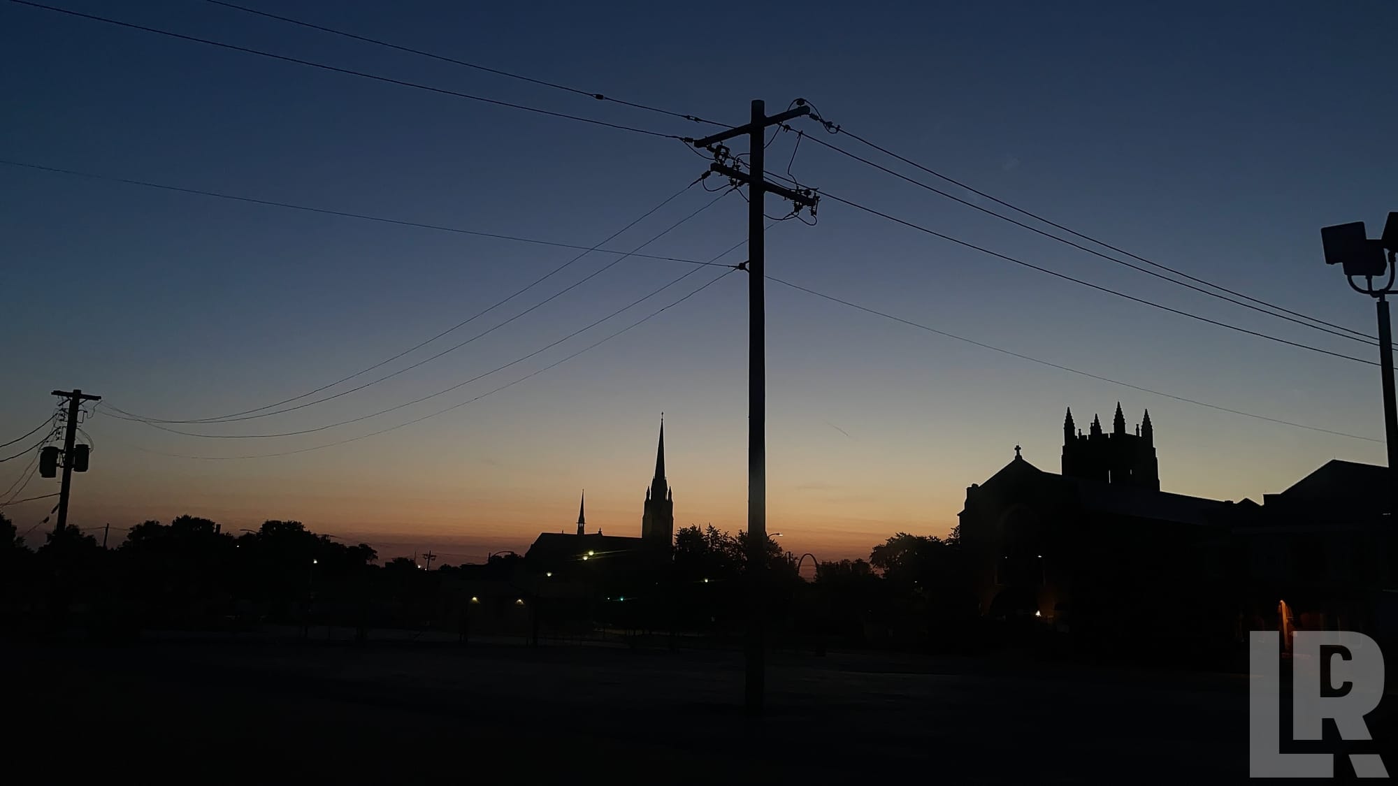 Silhouetted church steeples and buildings at dusk with power lines against a colorful sunset sky