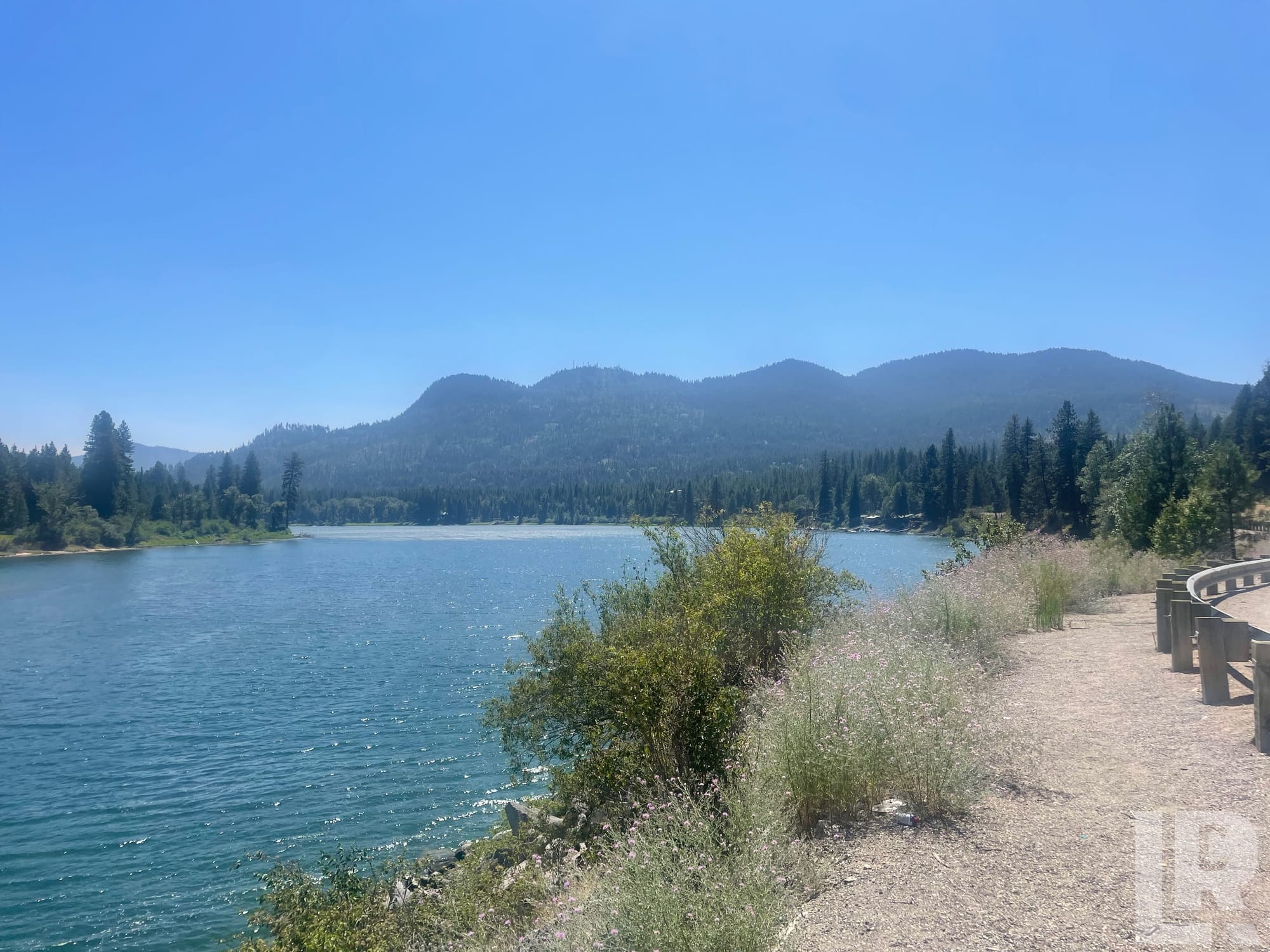 Turquoise lake beside a gravel road with forested mountains in the background on a sunny day