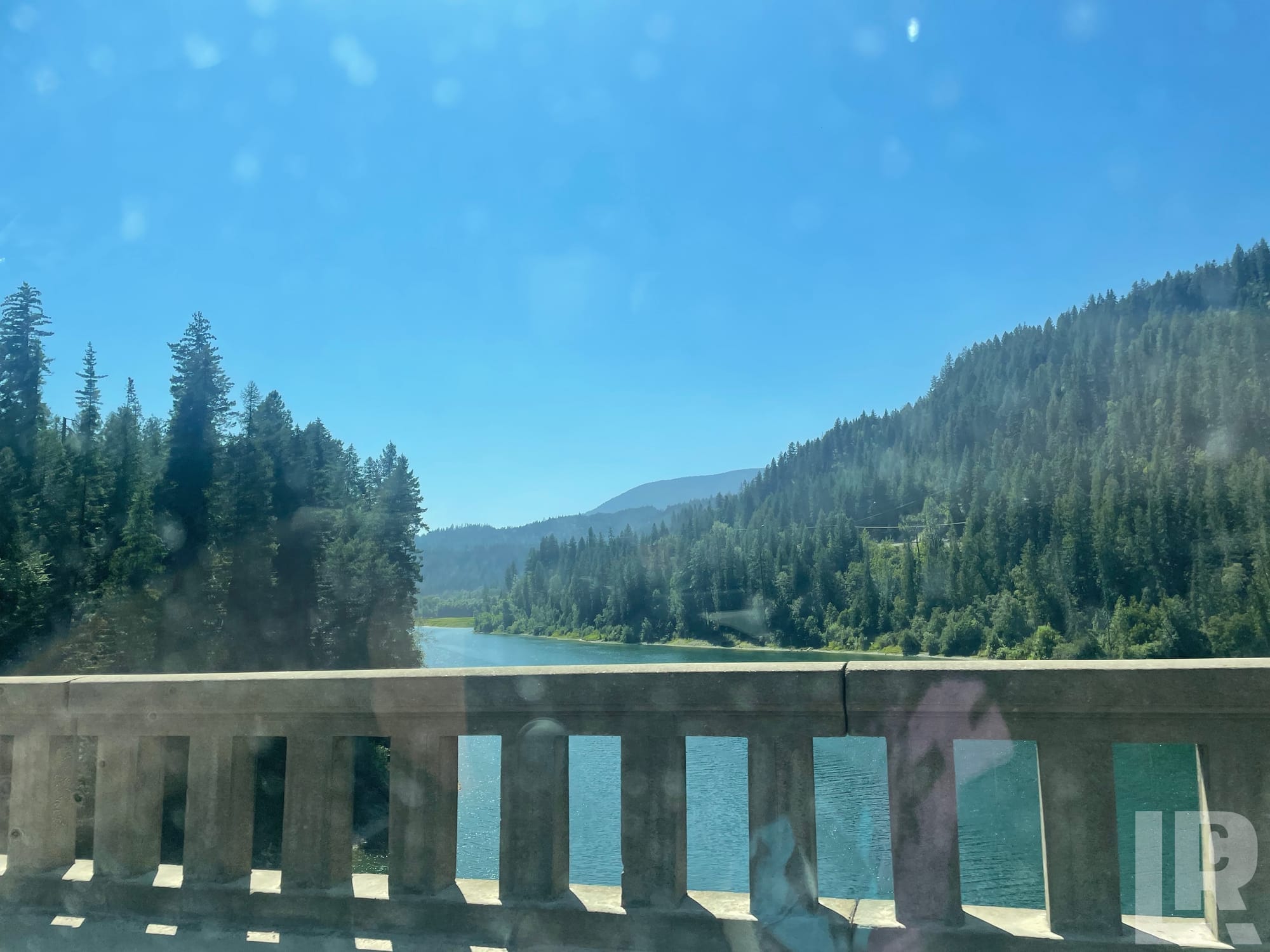 Bridge railing overlooking a turquoise river surrounded by pine-covered mountains on a sunny day