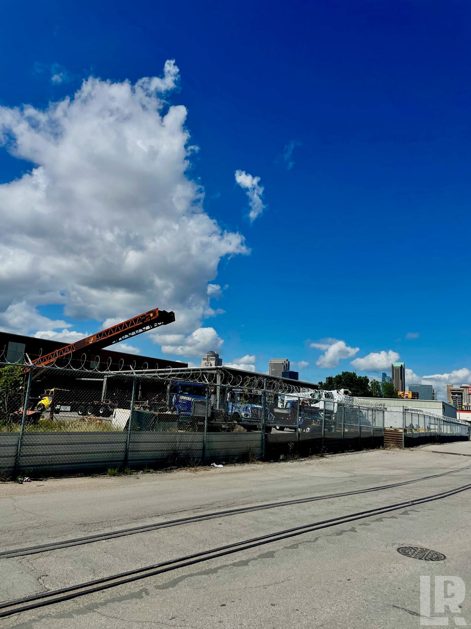 Razor-wire-topped chain-link fence surrounding an industrial truck yard with a crane boom extended, city skyline in the distance