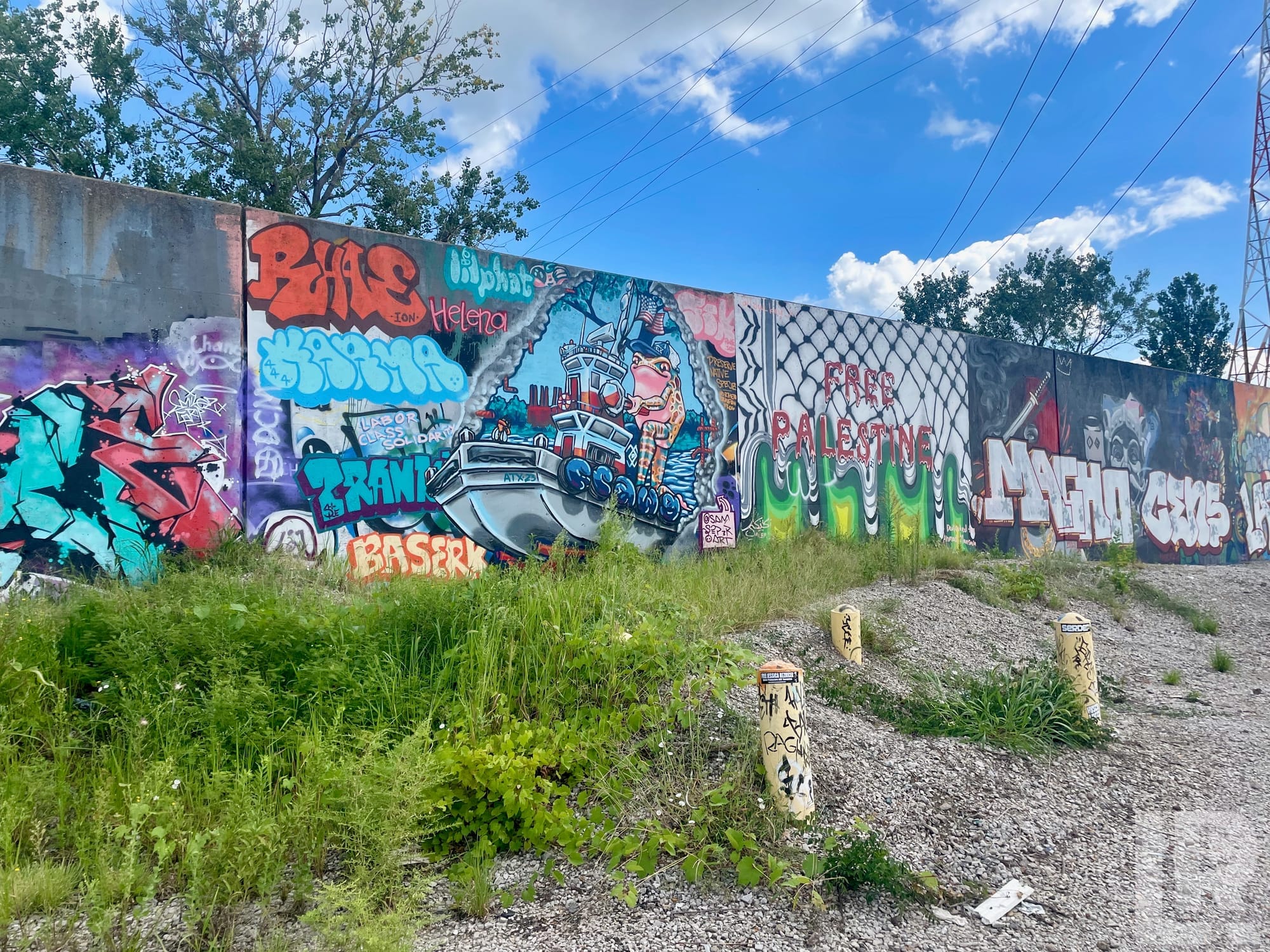 Graffiti-covered concrete wall with colorful murals and tags including a Free Palestine message, with weeds and bollards in the foreground