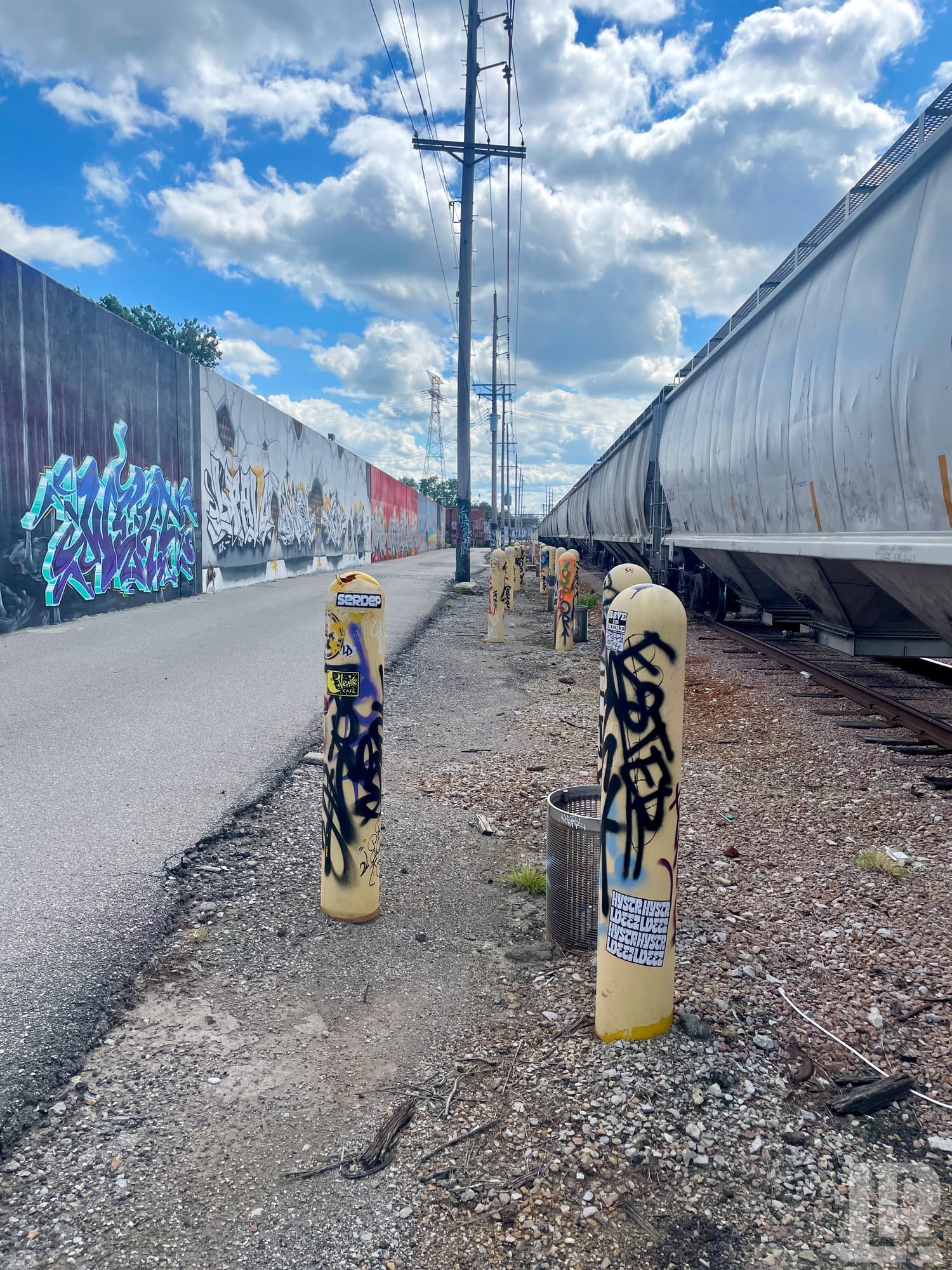Graffiti-tagged yellow bollards along a path between a mural wall and freight train cars