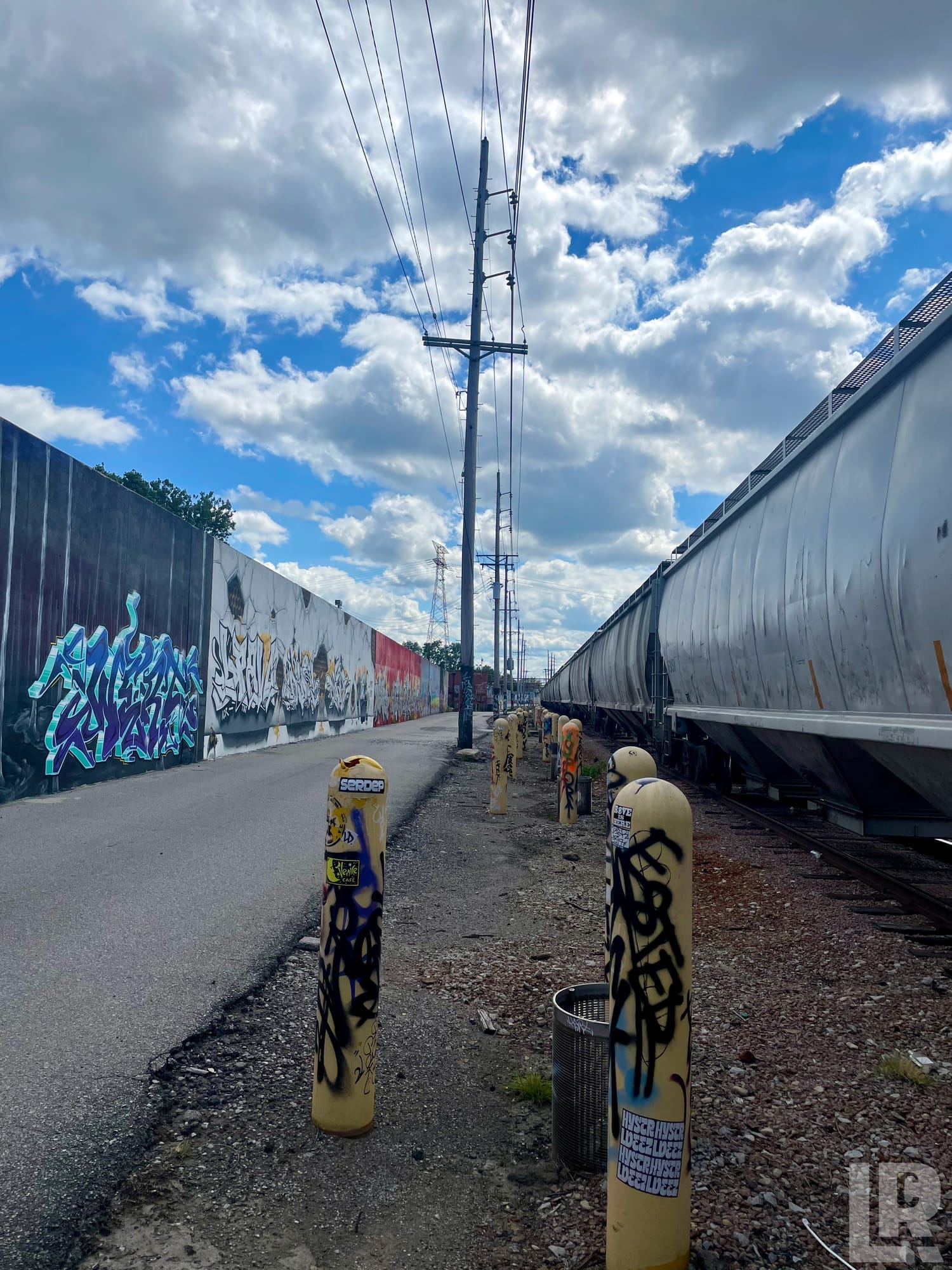 Graffiti-covered wall alongside a freight train and yellow bollards on an urban rail corridor path