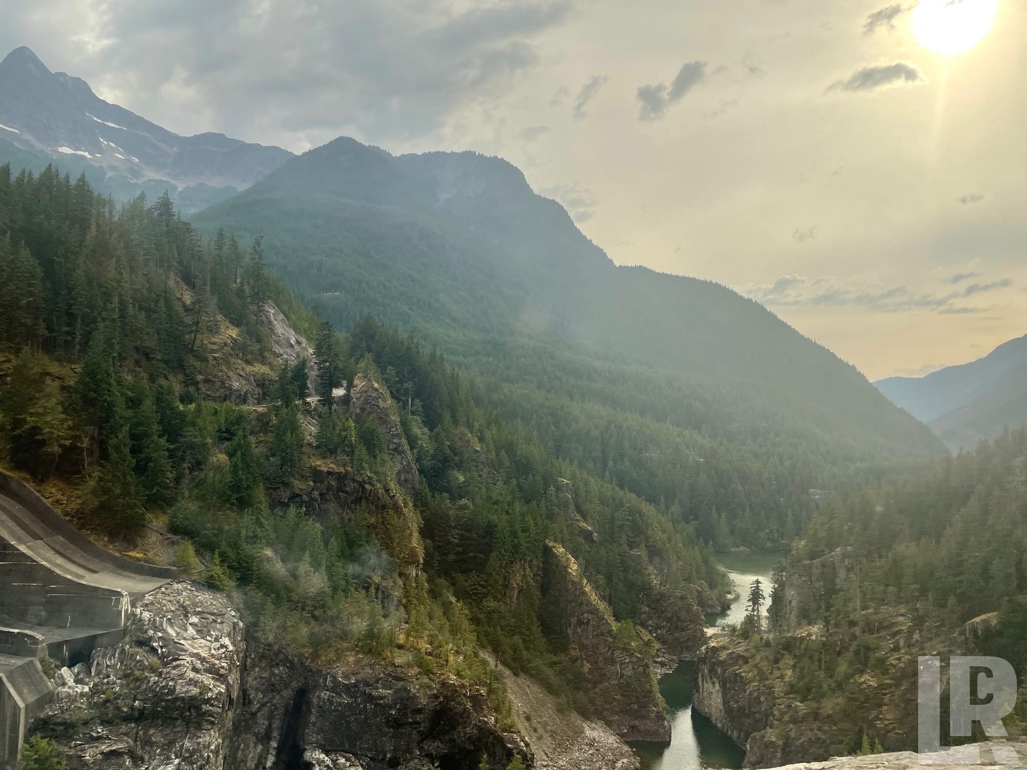 Mountain valley with evergreen forests, rocky cliffs, a river below, and a dam in the foreground on a hazy day