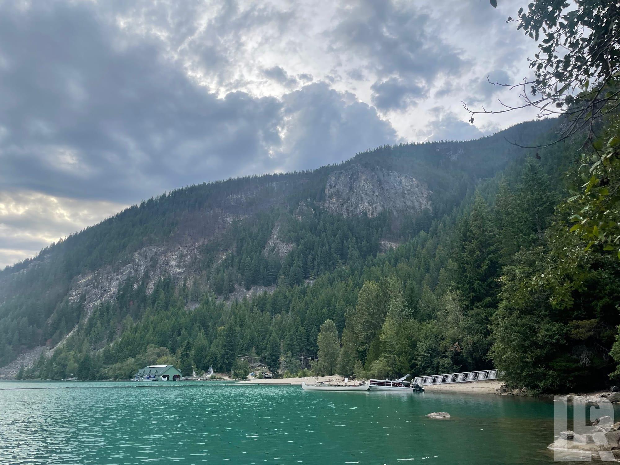Turquoise mountain lake with forested rocky hillside, sandy beach, and docked boat under cloudy sky