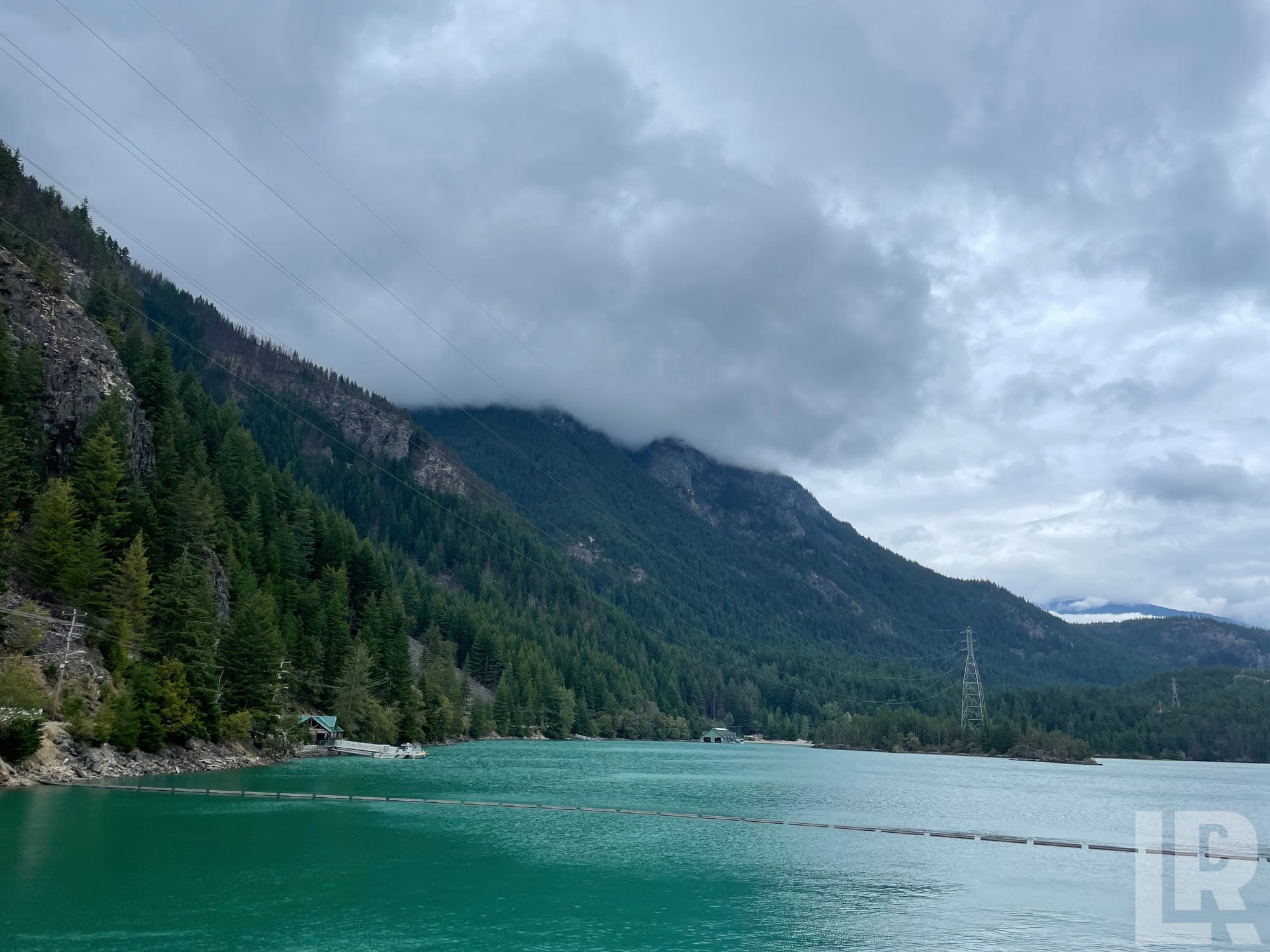 Turquoise mountain reservoir flanked by conifer-covered slopes under overcast skies with power lines overhead