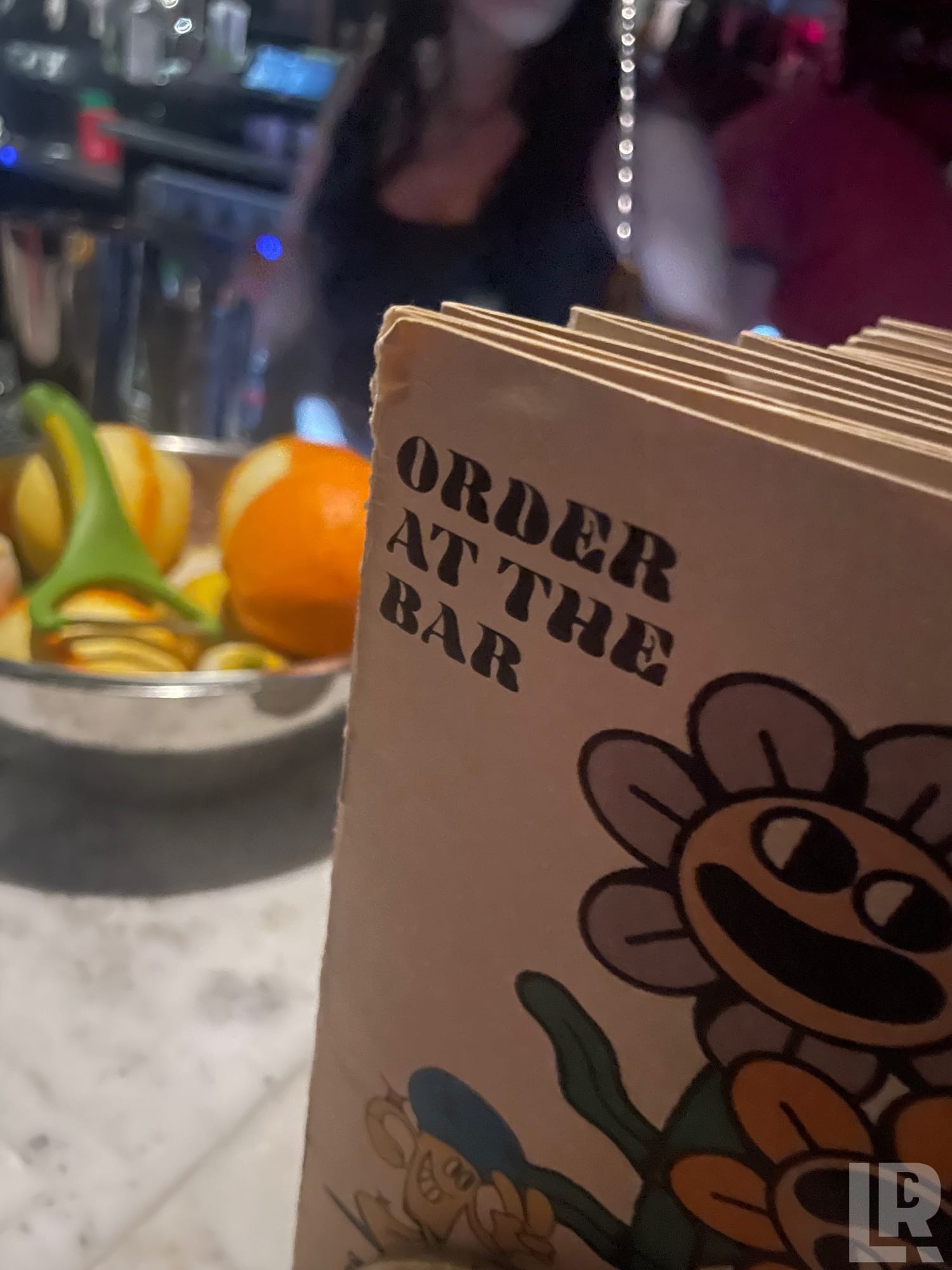 Bar menus reading 'Order at the Bar' with cartoon flower art, beside a bowl of orange and lemon slices on a marble bar top